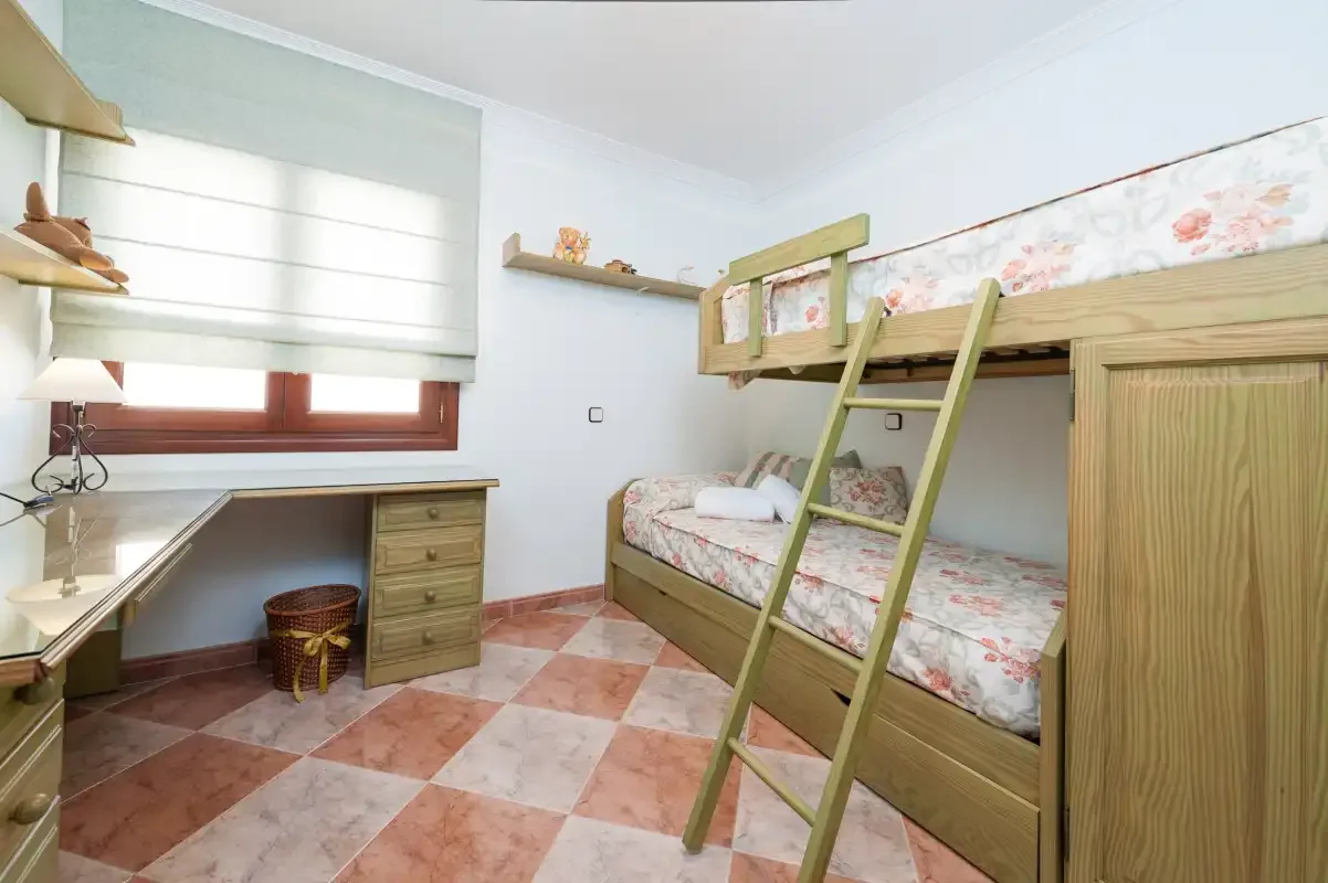 A bedroom with a wooden bunk bed, floral bedding, and a desk beneath the window.