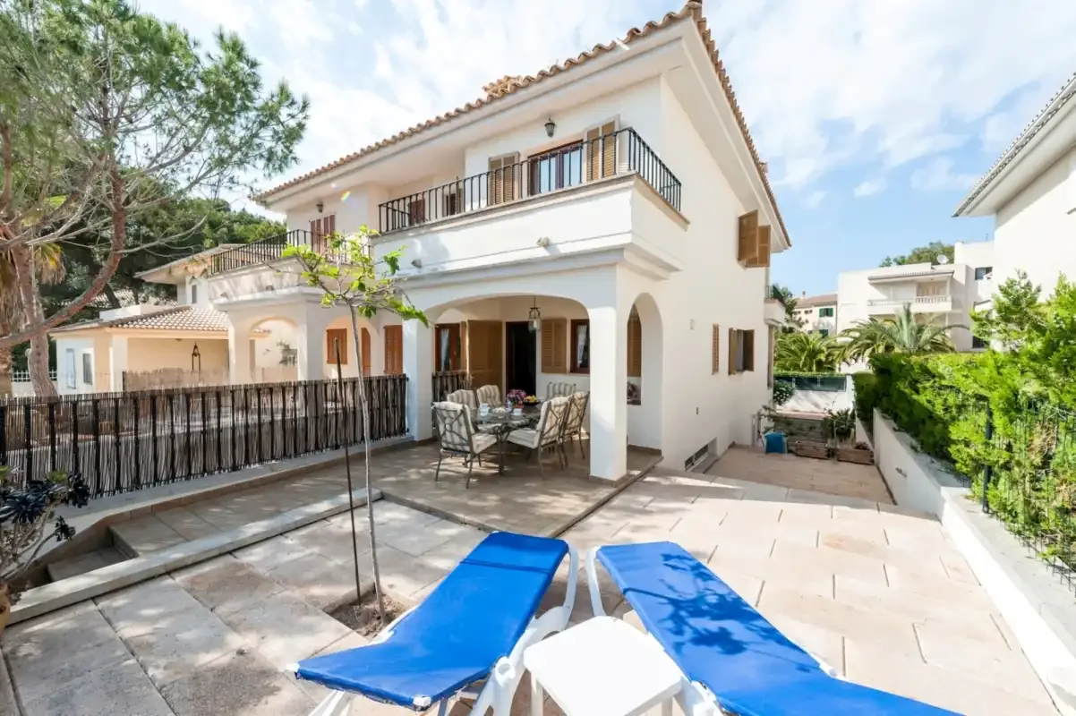 A white two-story house overlooks a paved patio with outdoor seating and loungers.