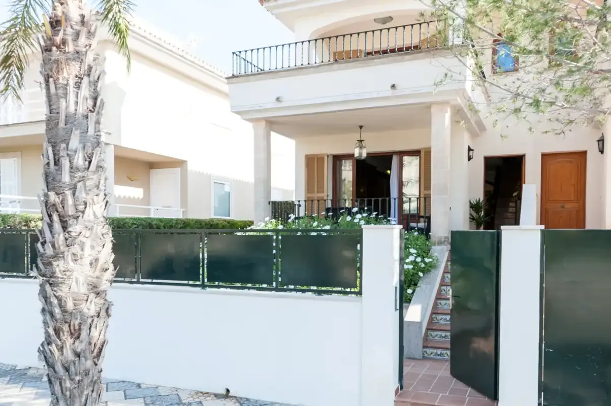A white multi-level house stands behind a low front wall and open gate.