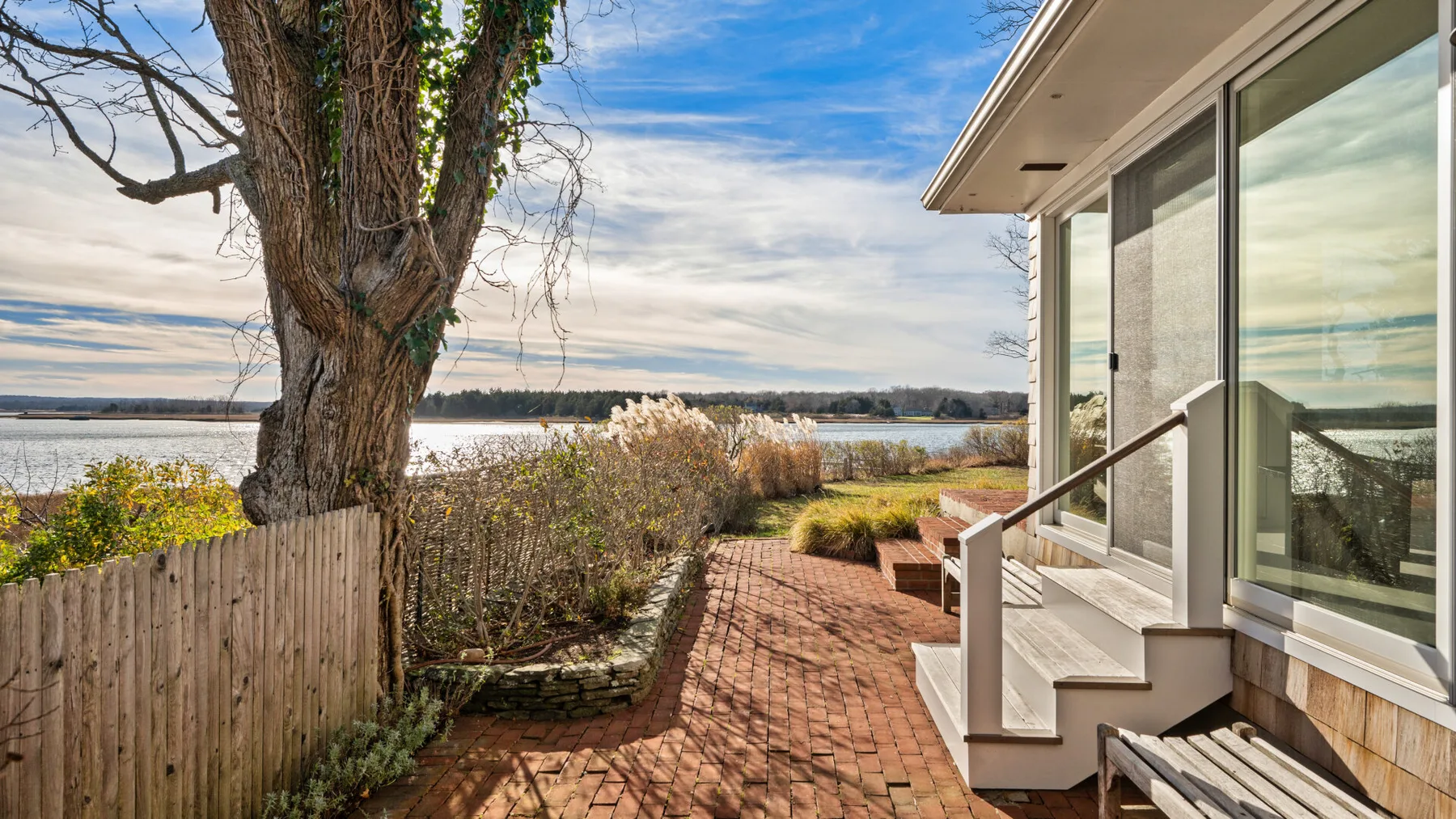 Un patio de ladrillo junto a una casa moderna tiene vistas a un lago y la vegetación circundante.