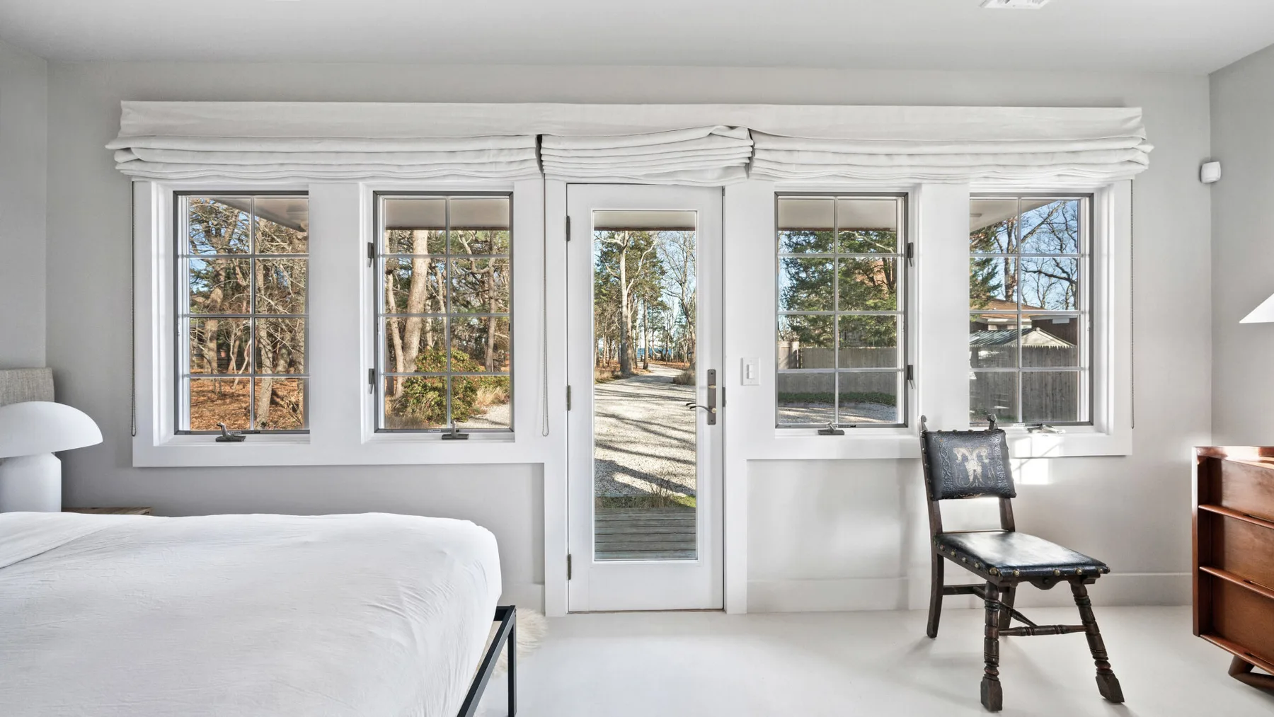 Un dormitorio luminoso tiene una cama junto a ventanas y una puerta de vidrio con vista a los árboles.