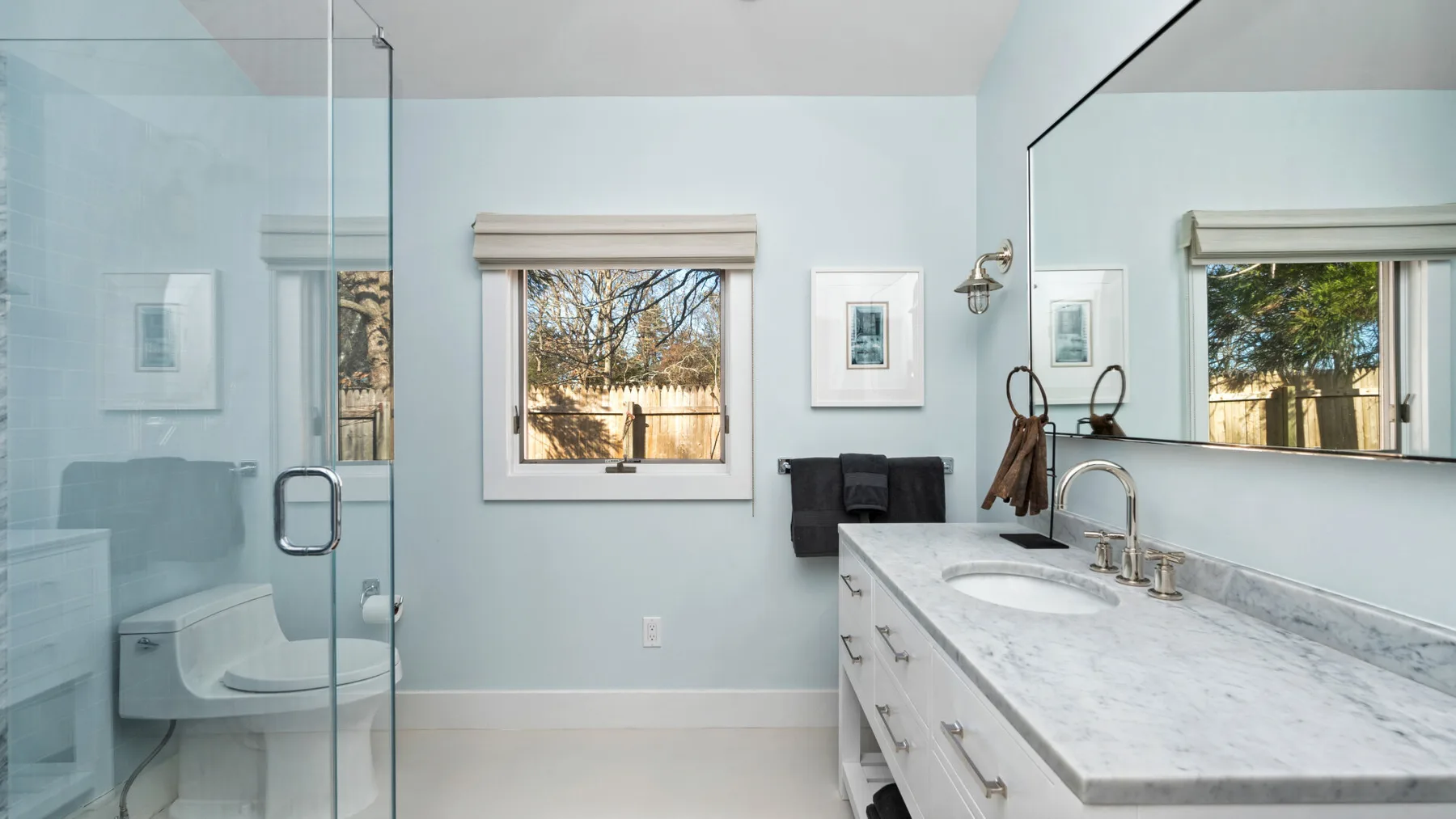 Un baño luminoso tiene una ducha de vidrio, un mueble de lavabo largo y una ventana.