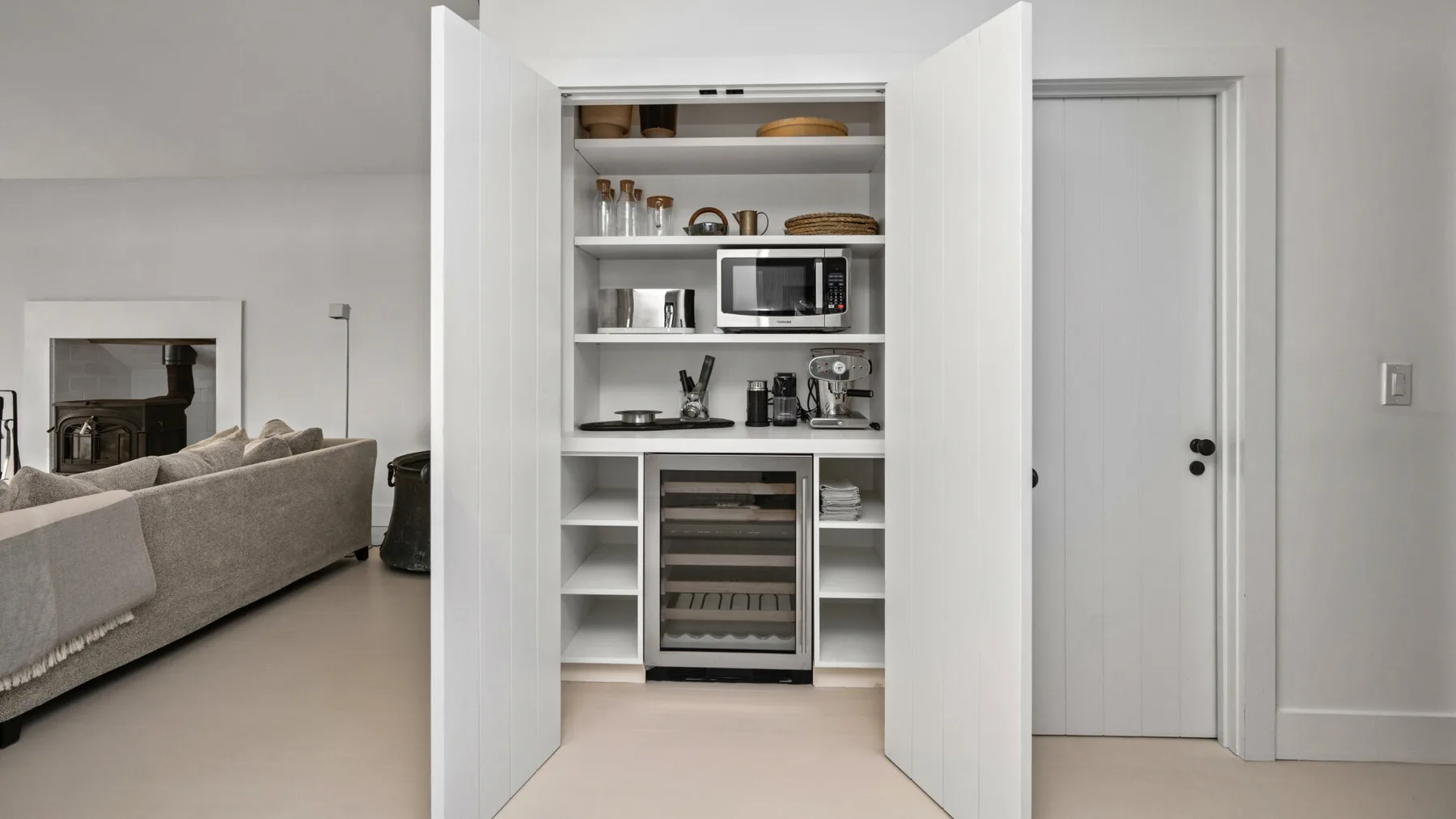 Un armario de cocina abierto muestra vajilla y electrodomésticos junto a una sala de estar.