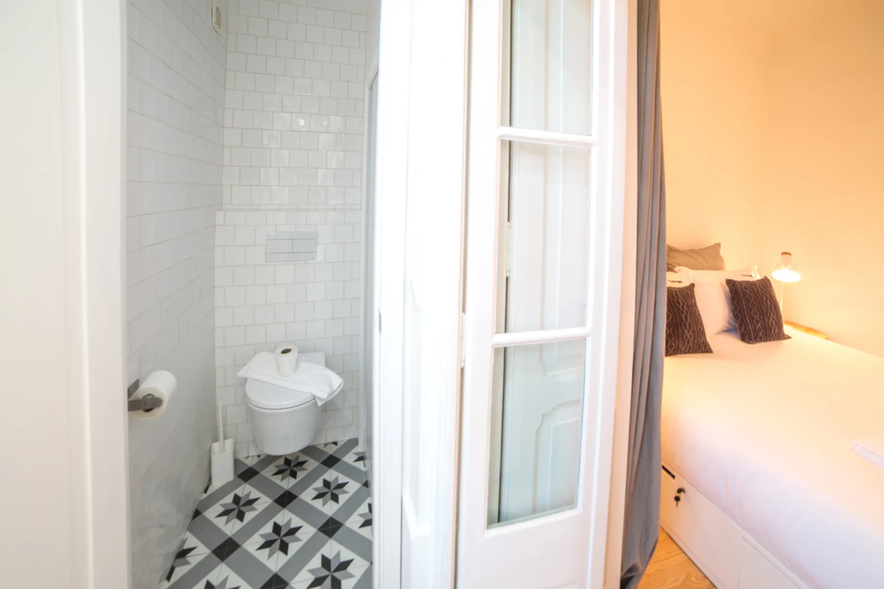 A bathroom with patterned floor tiles opens onto a bedroom with a bed.