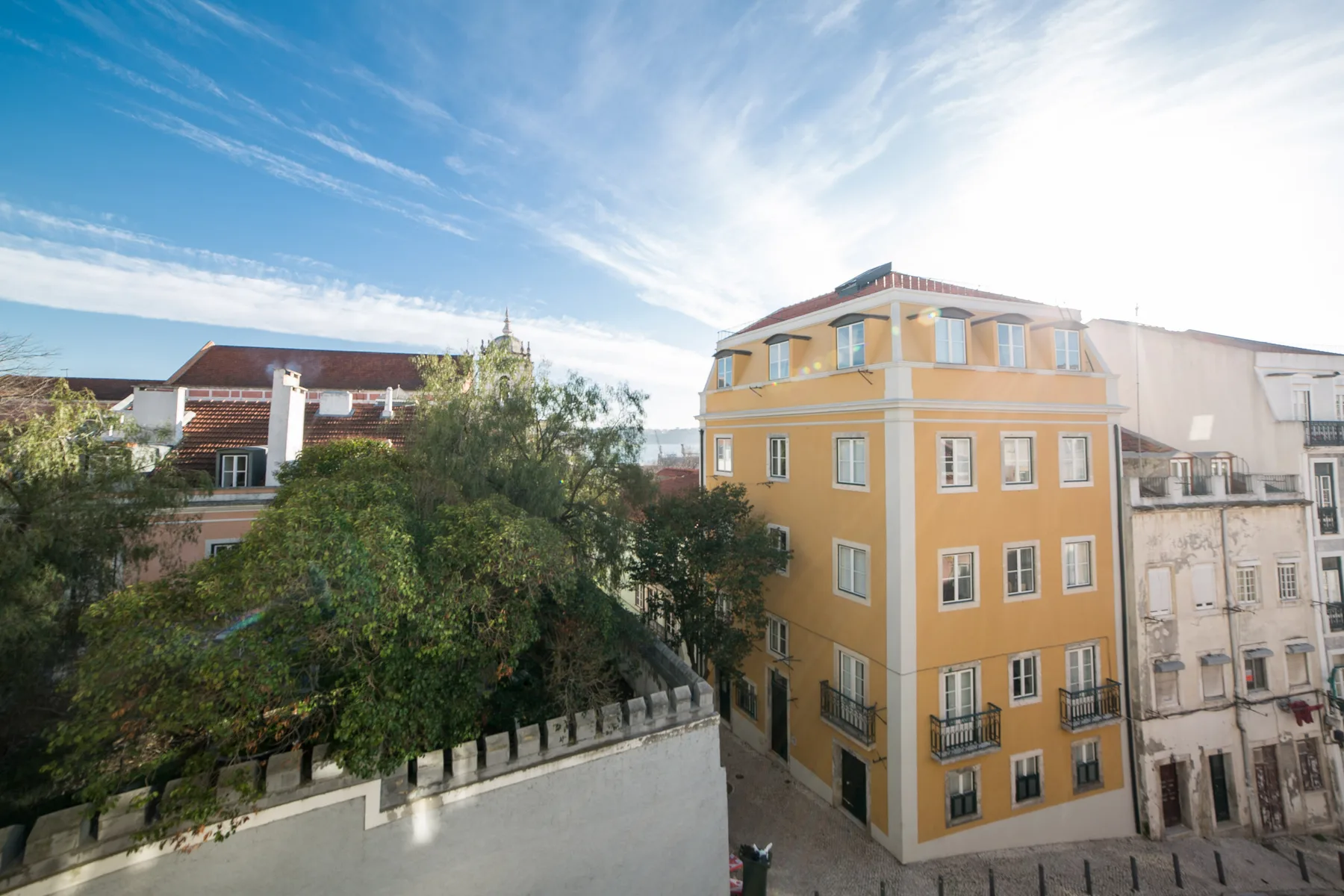 A sunny urban streetscape with an ochre building and leafy trees.