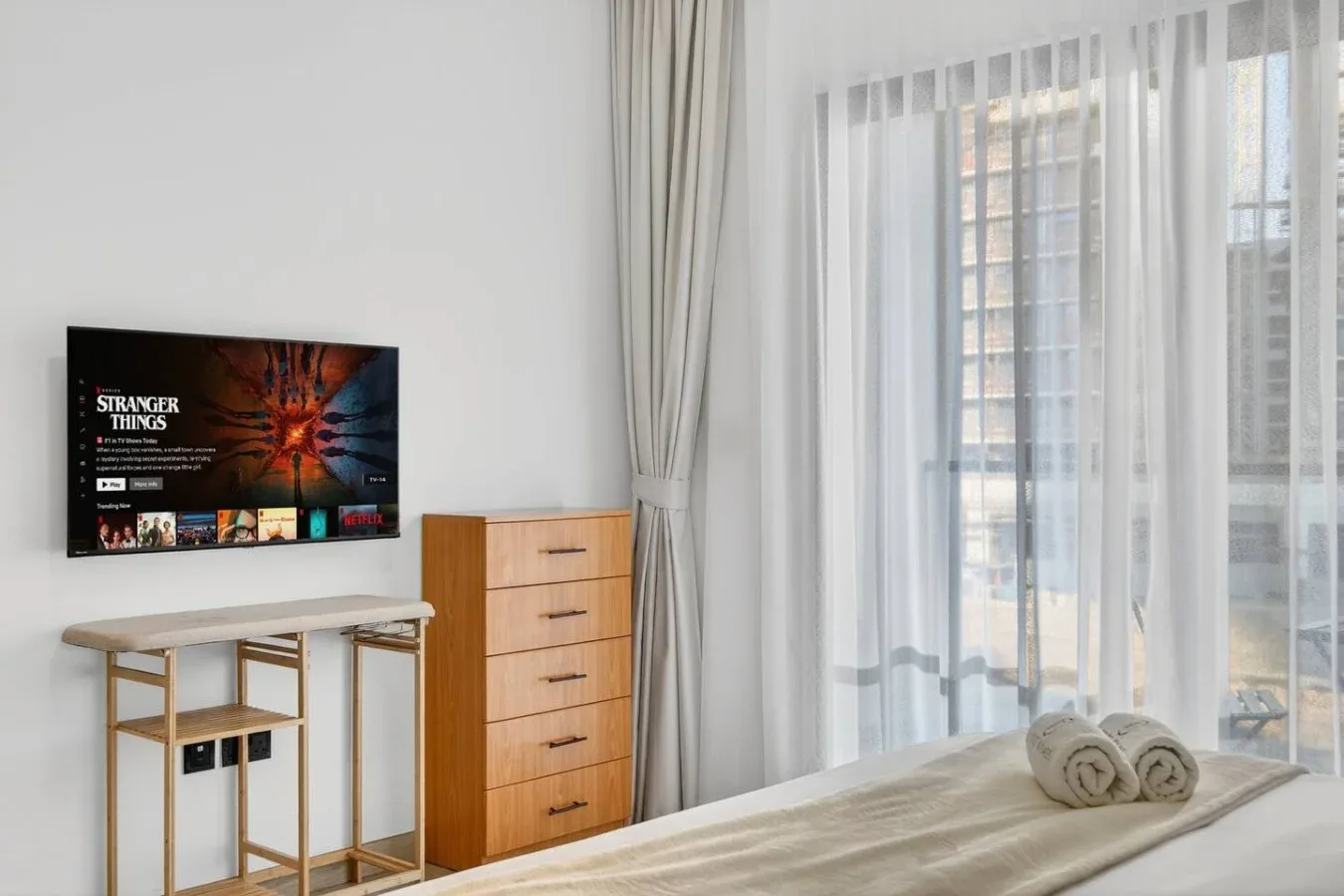 Un dormitorio luminoso con televisor, cómoda y cortinas translúcidas junto a la ventana.