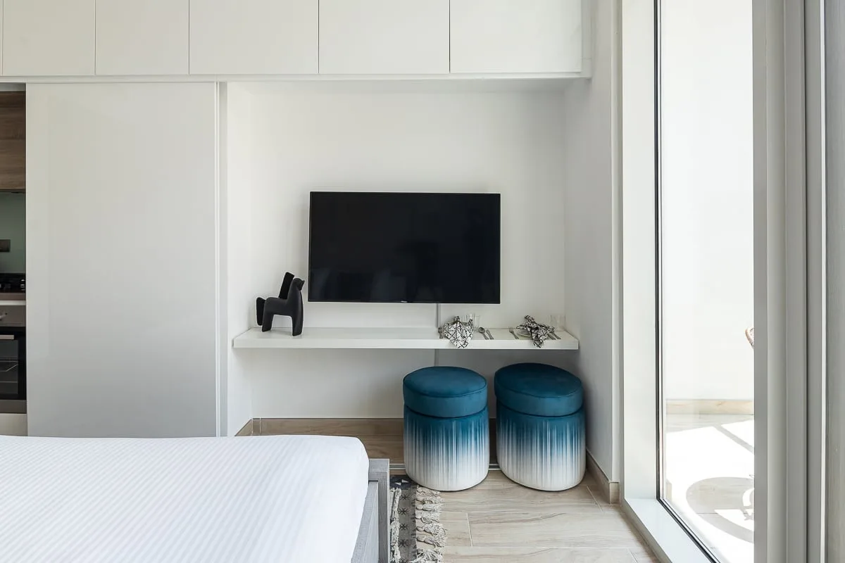A bright bedroom with a television above a built-in desk and two stools.