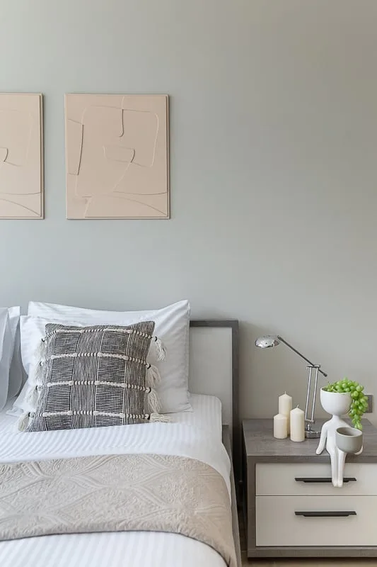 A neatly made bed sits beside a small bedside table beneath framed artwork.