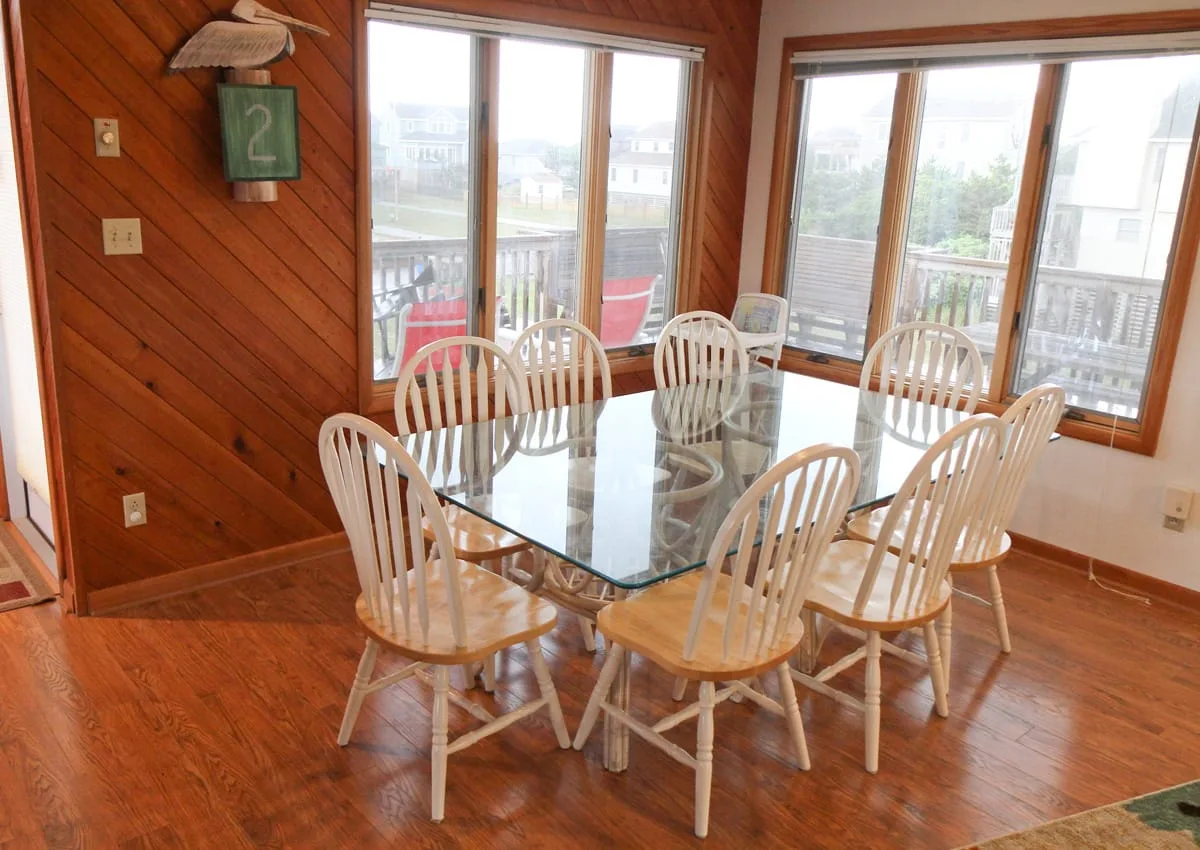 Comedor con una mesa de vidrio y asientos de madera junto a grandes ventanas.