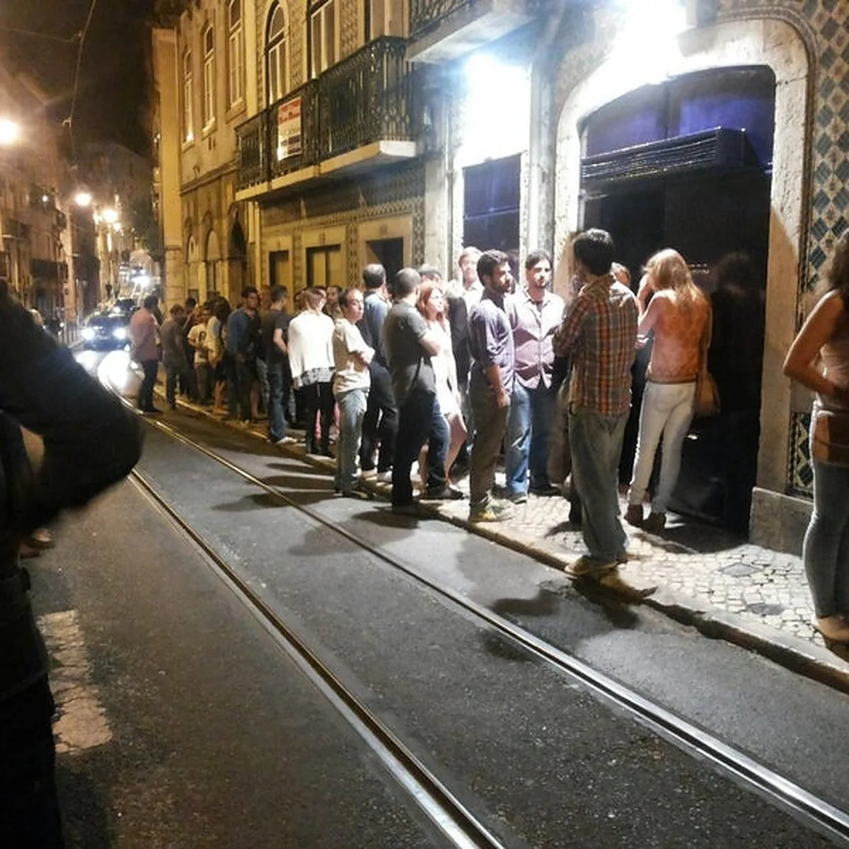 People gather along a city street beside tram tracks at night.