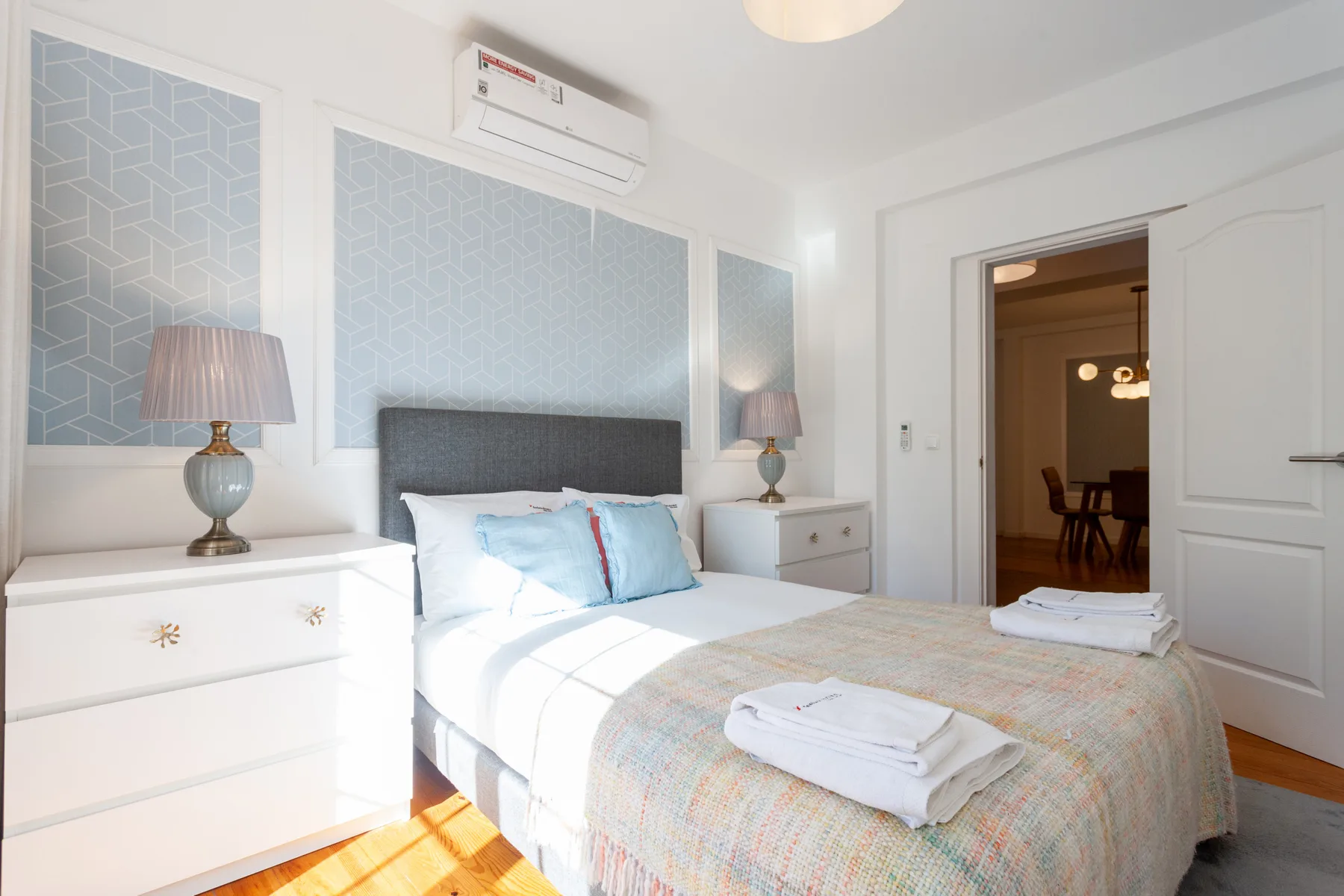 A bright bedroom with a made bed, bedside tables, and pale patterned wall panels.