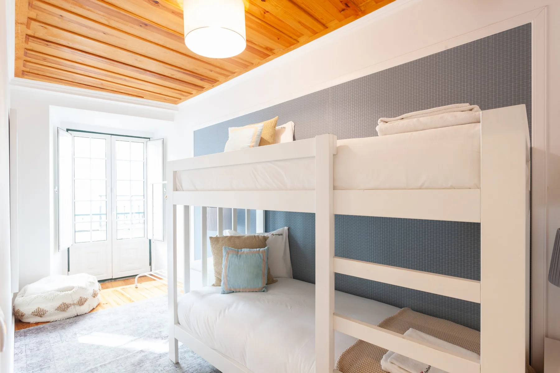 A bright bedroom with bunk beds, a window, and a wood-paneled ceiling.