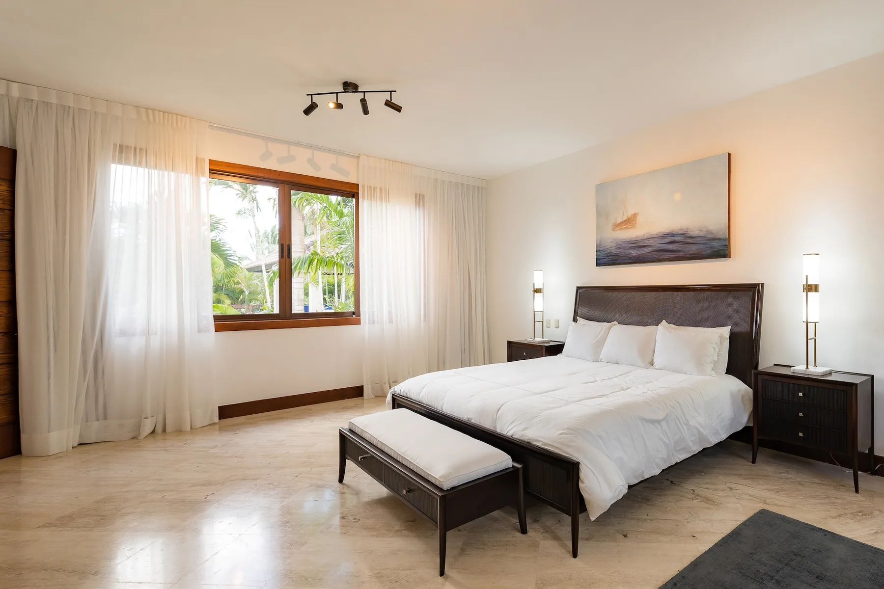 A bright bedroom with a large bed and a window framed by sheer curtains.