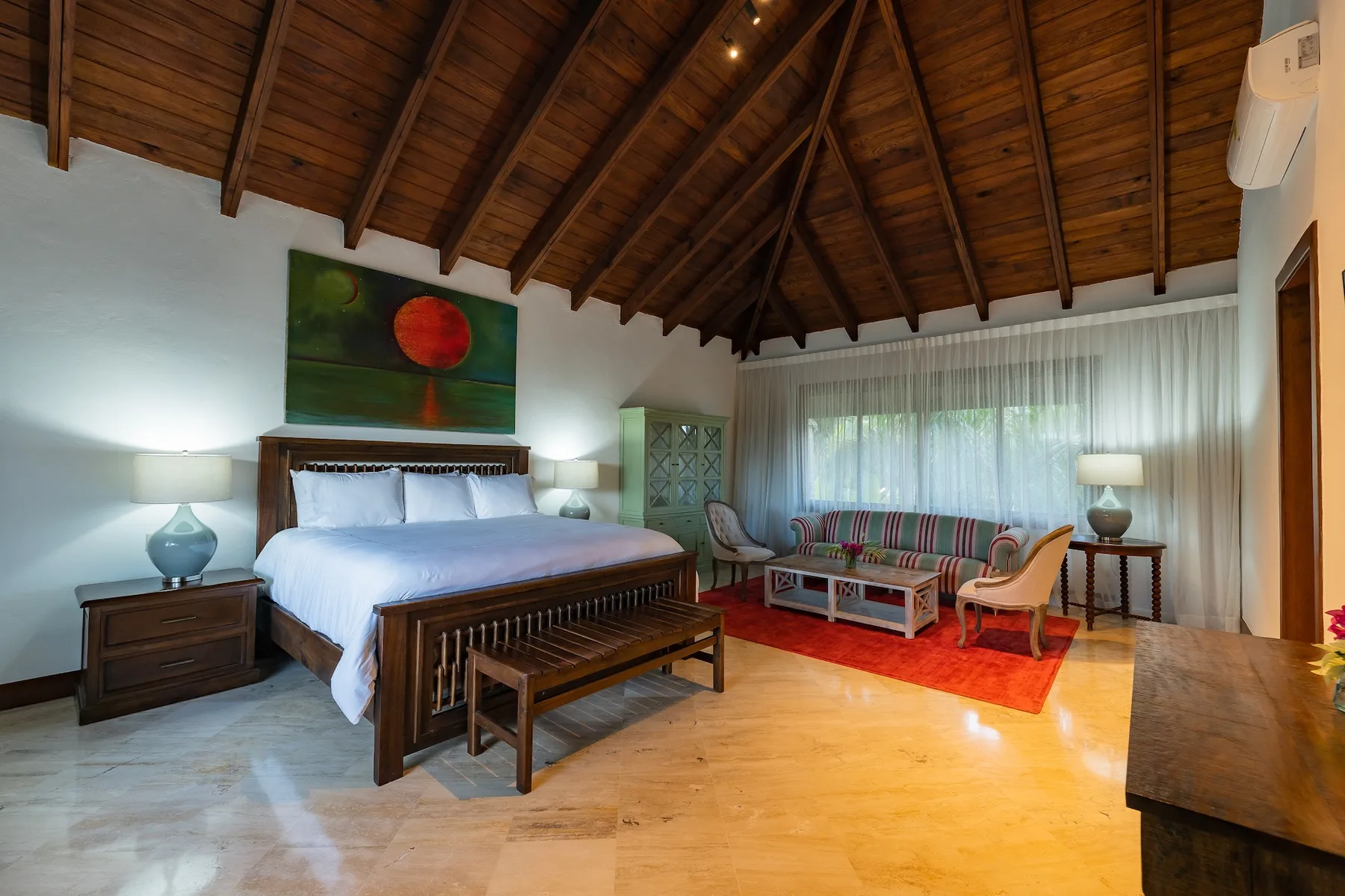 A bedroom with a large bed and a sitting area beneath a wooden vaulted ceiling.
