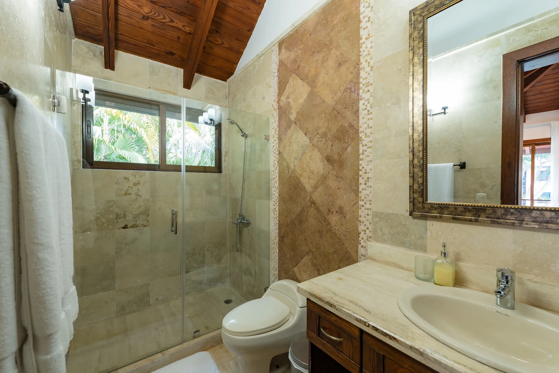 A bathroom with a glass shower, toilet, and sink beneath a wooden ceiling.