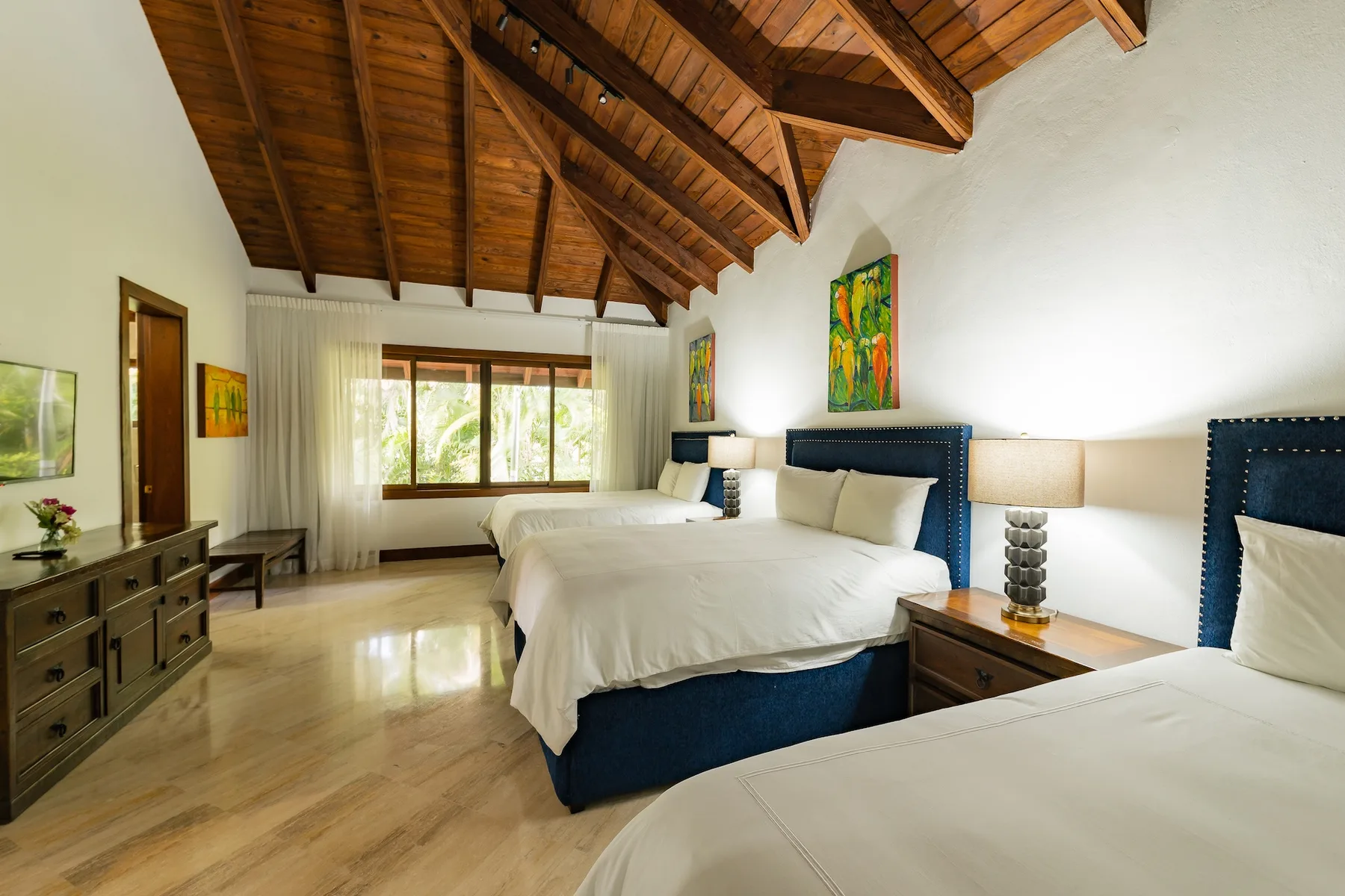 A bright bedroom with beds, a wooden ceiling, and a large window.