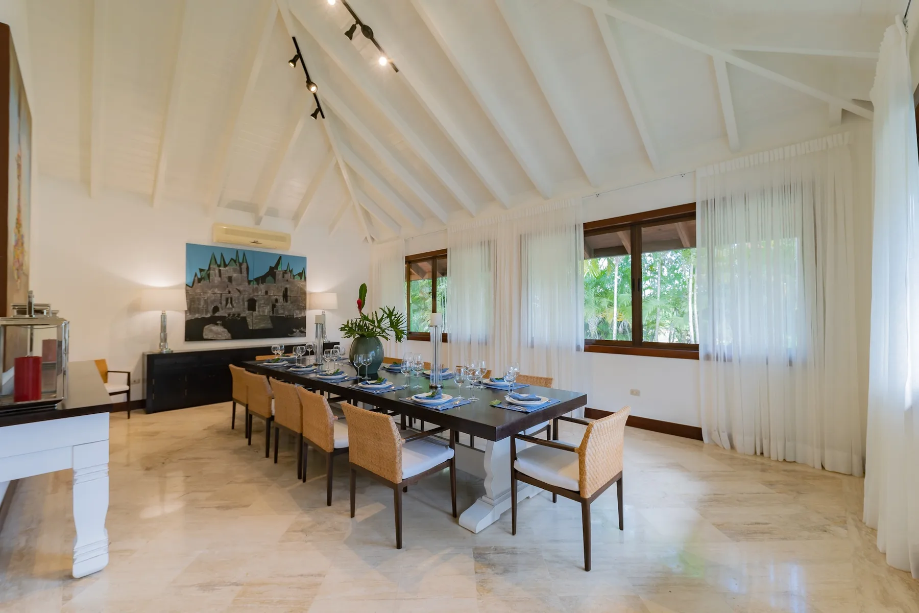 A dining room with a long table set beside large windows.