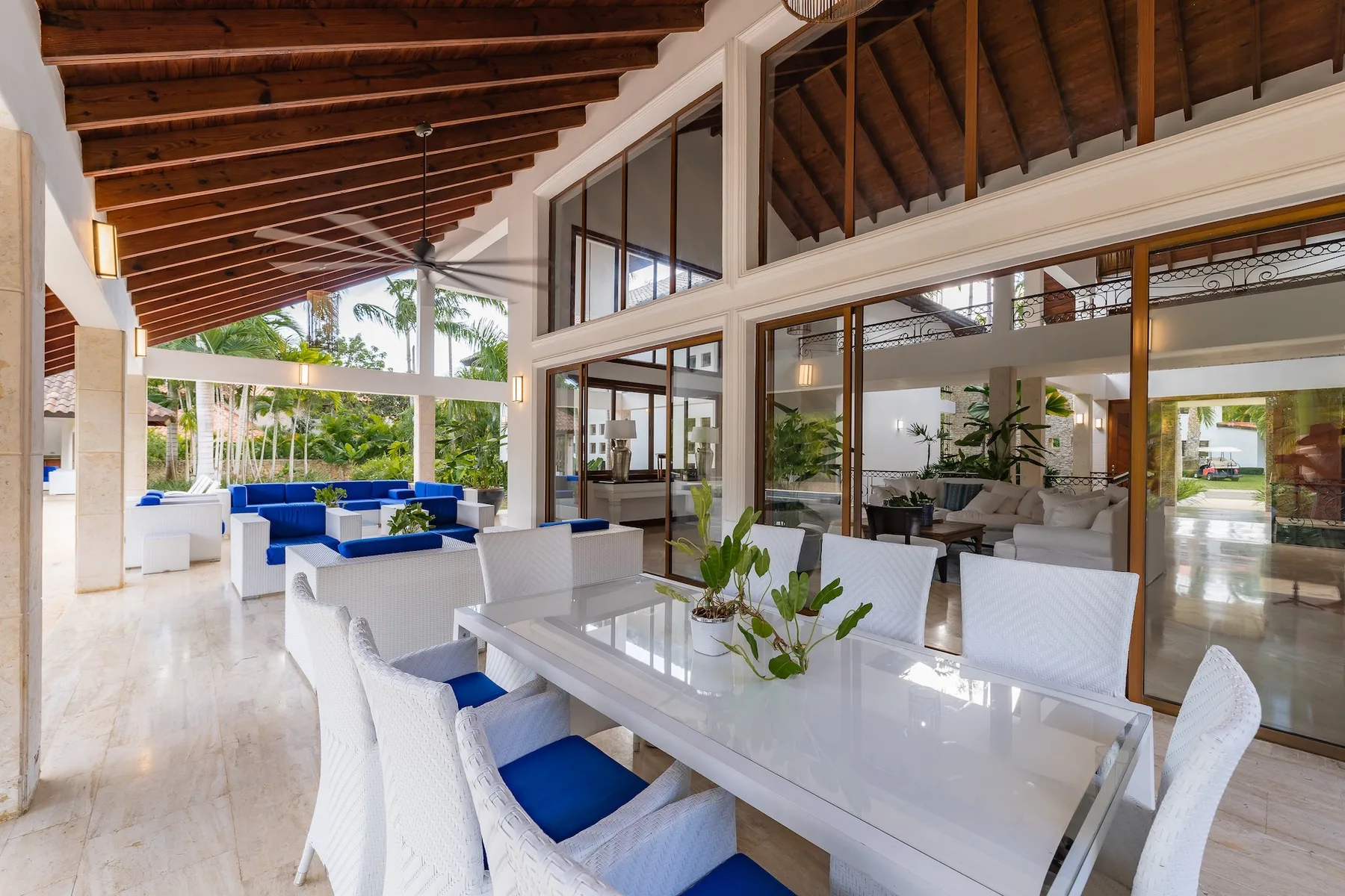 A covered outdoor dining area with white seating and a long table beside glass walls.