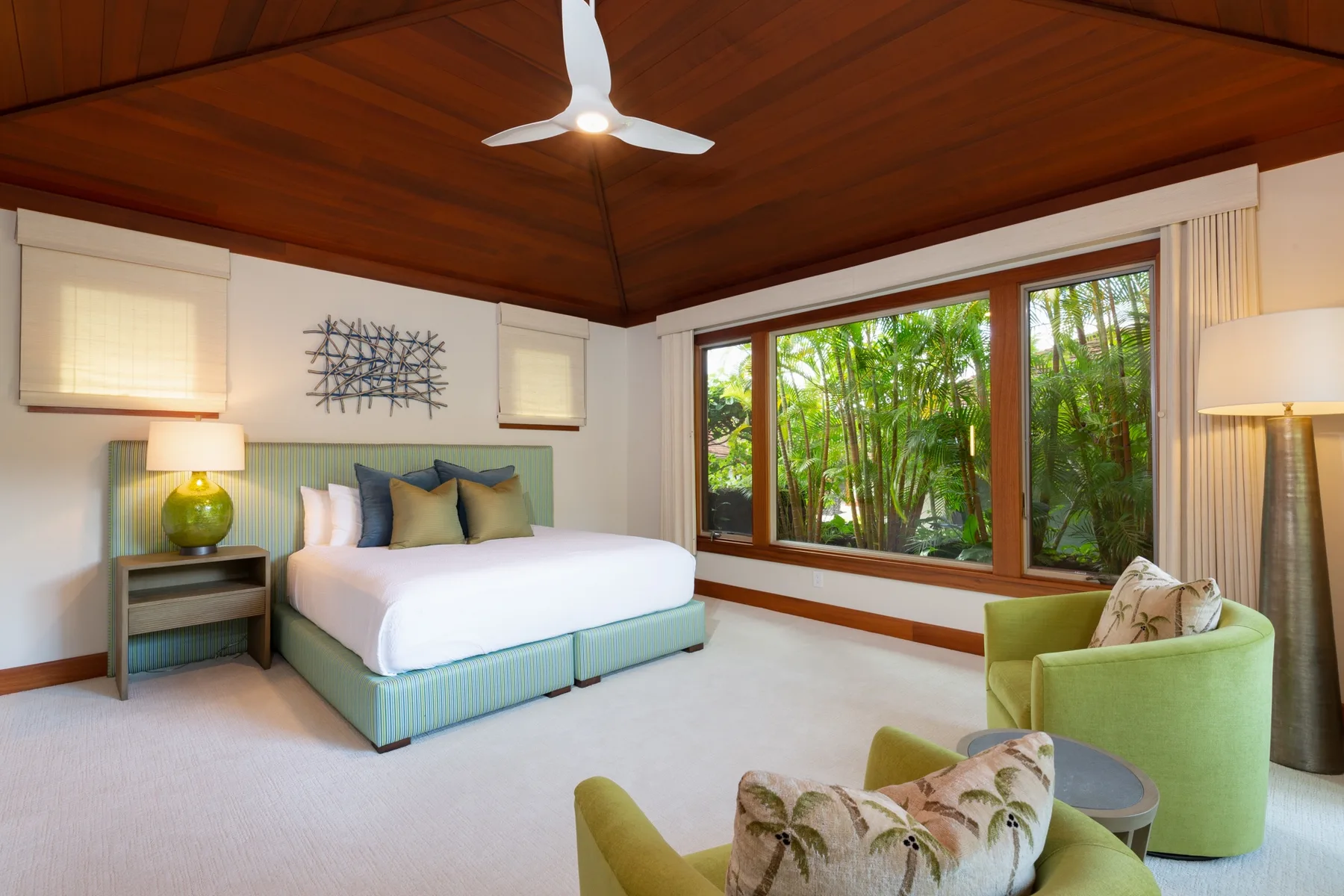 A bedroom with a bed, green seating, and large windows overlooking leafy greenery.