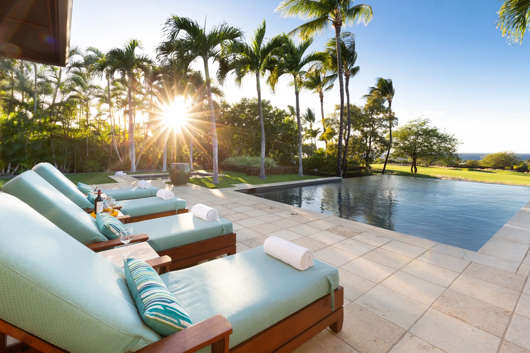 Outdoor pool beside cushioned seating, palm trees, and a lawn.
