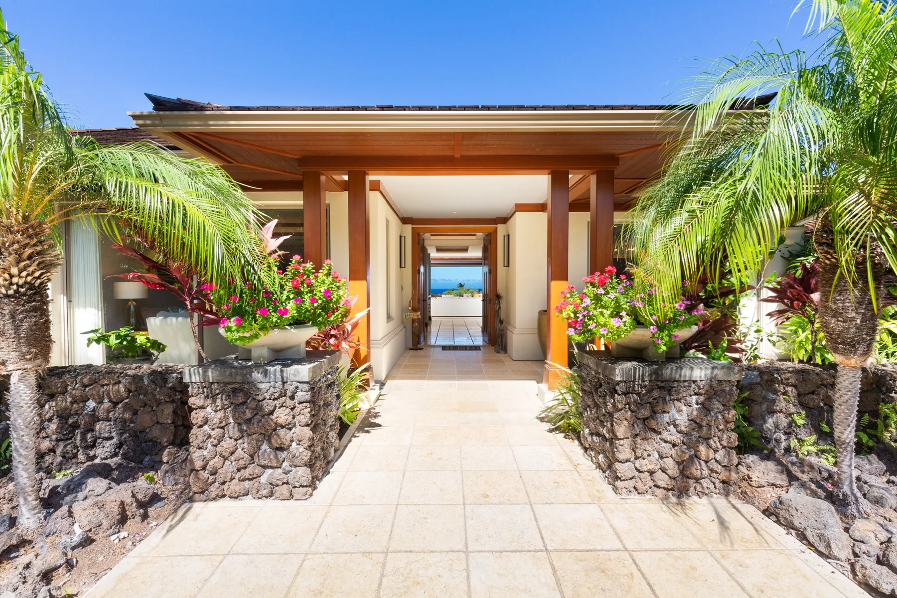 A sunlit building entrance opens onto a tiled walkway, framed by palms and flowering plants.