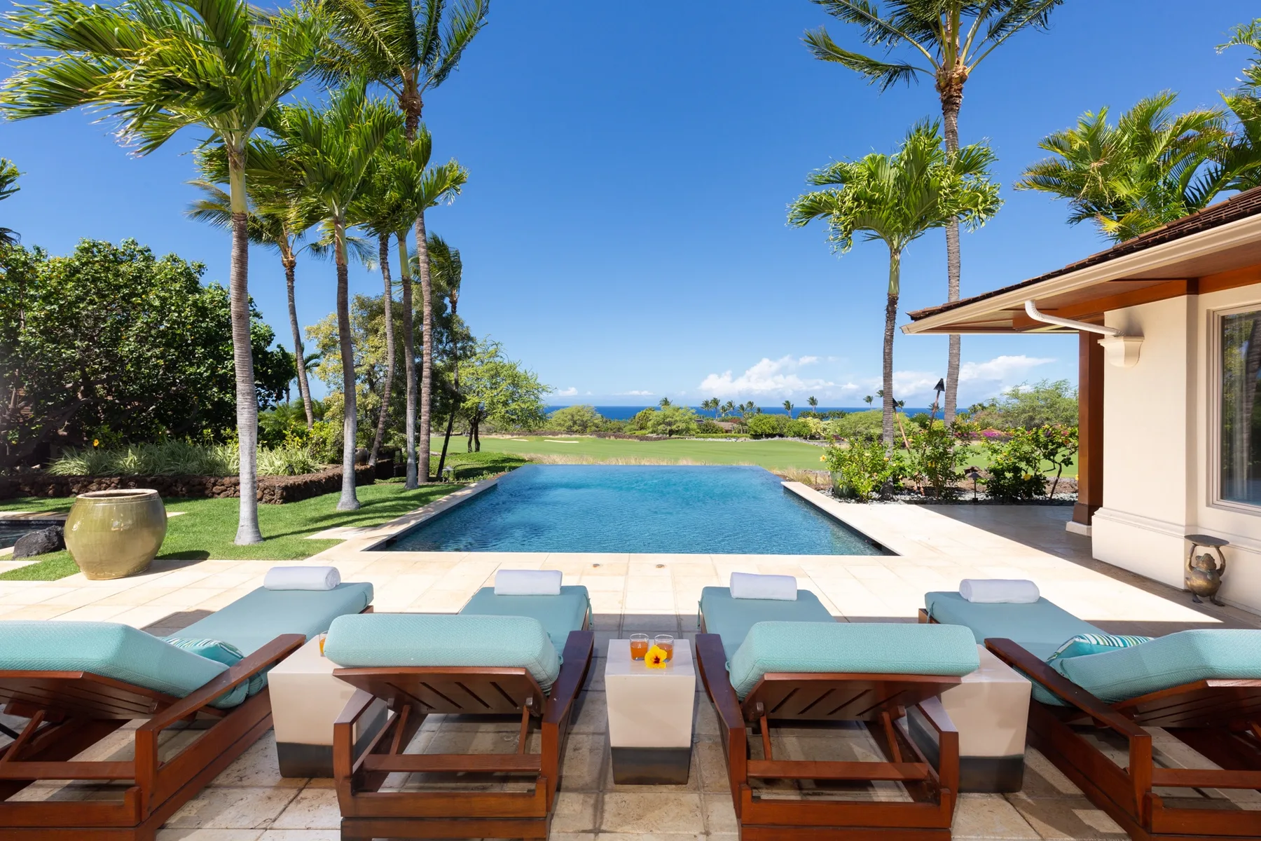A swimming pool beside a row of lounge seating, with palm trees and ocean views.