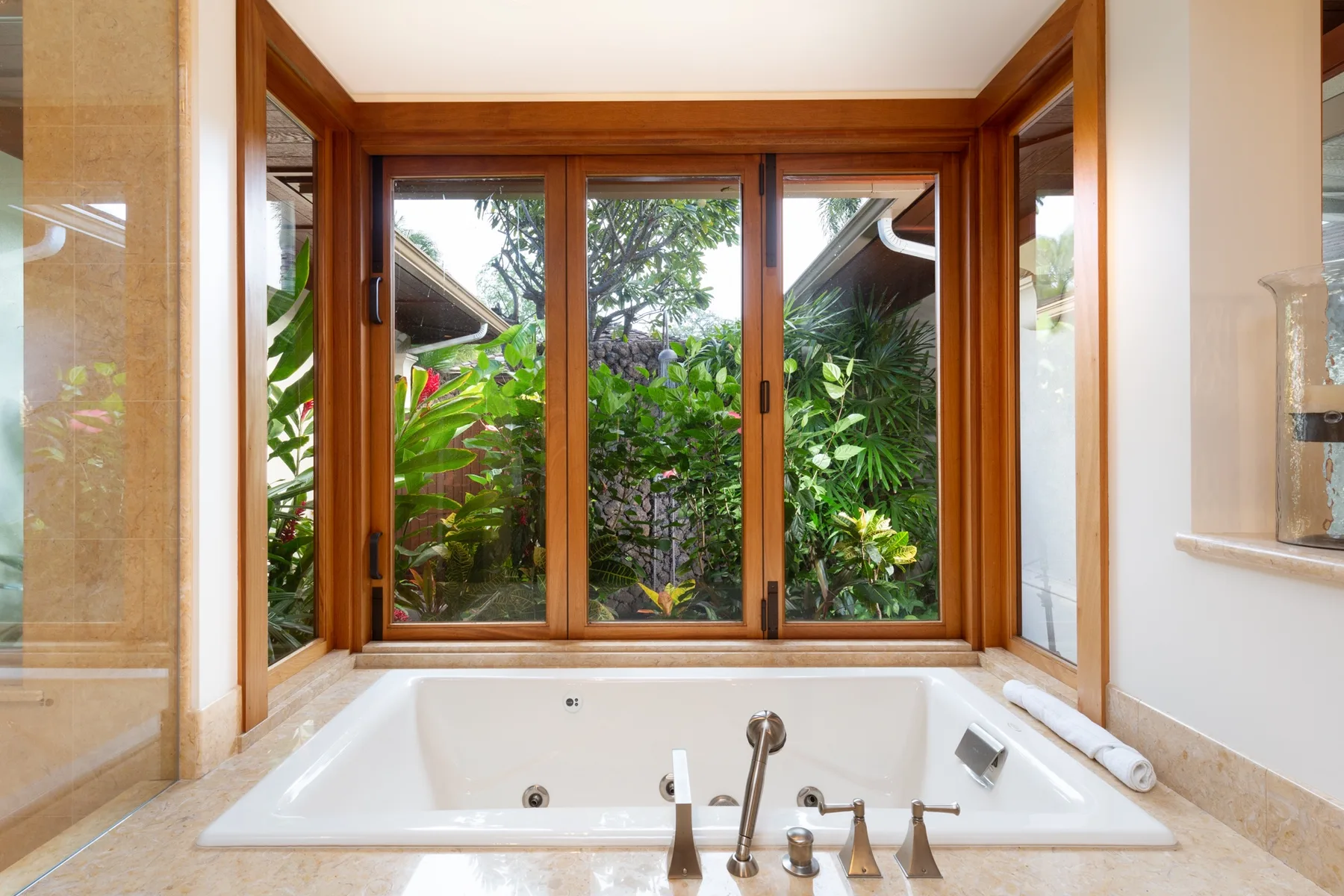 A bathtub sits beneath wood-framed windows overlooking lush greenery.