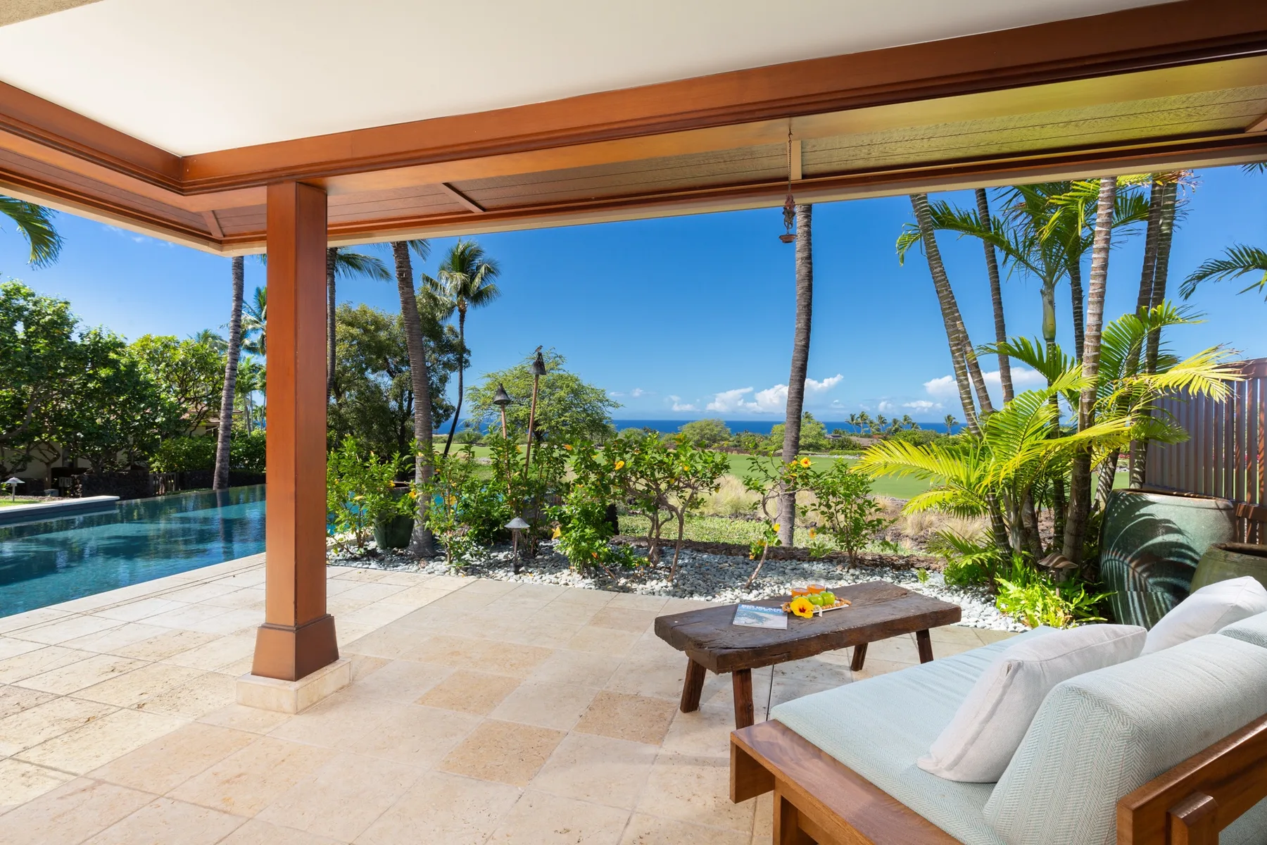 A covered patio with outdoor seating overlooks a pool, tropical landscaping, and the ocean.