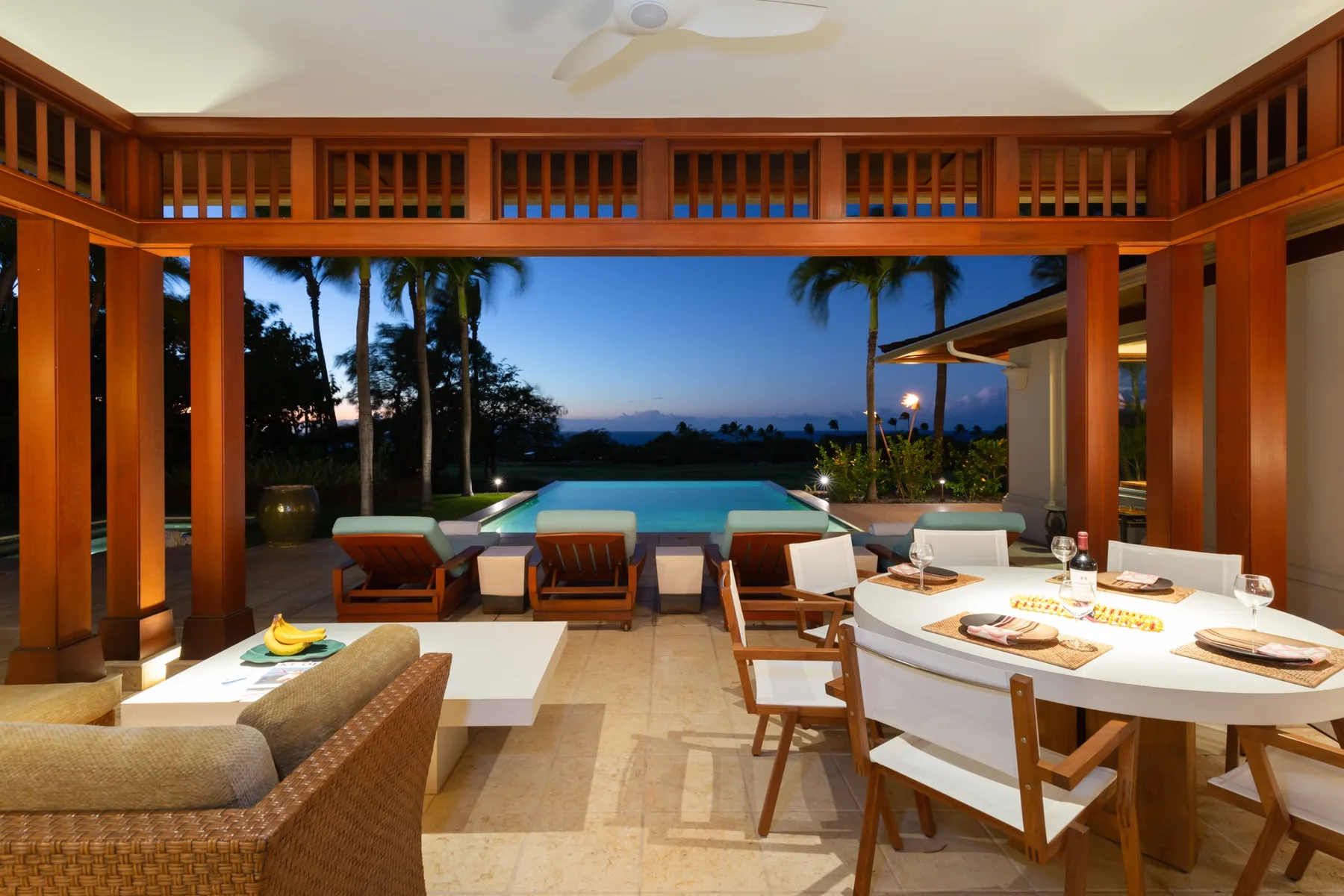 A covered patio with a dining table overlooks a pool and landscaped grounds at dusk.