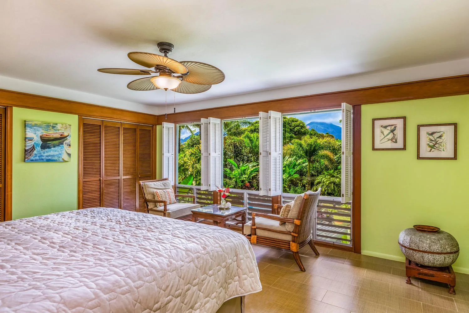 Un dormitorio con una cama y asientos junto a ventanas abiertas con vistas a abundante vegetación.