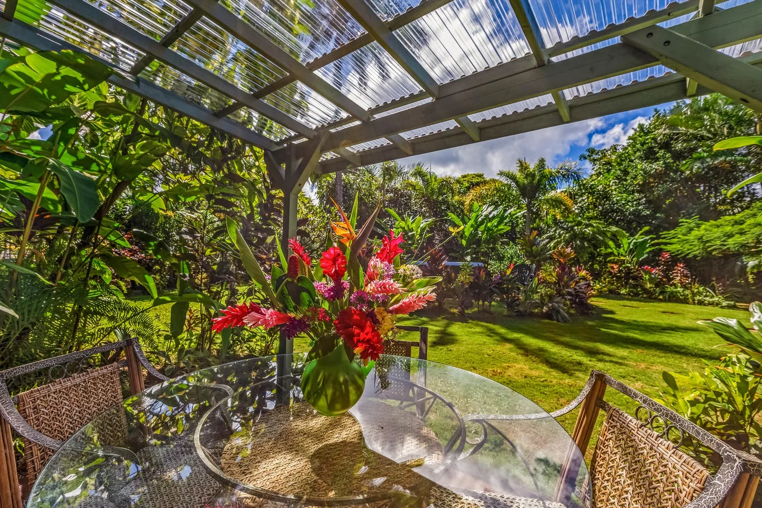 Un patio al aire libre con una mesa de vidrio y abundante vegetación.