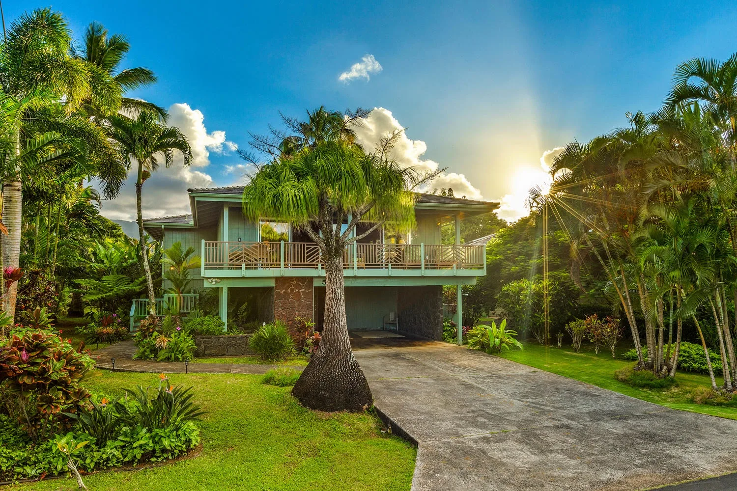 Una casa tropical con balcón está rodeada de palmeras, junto a una entrada para vehículos.