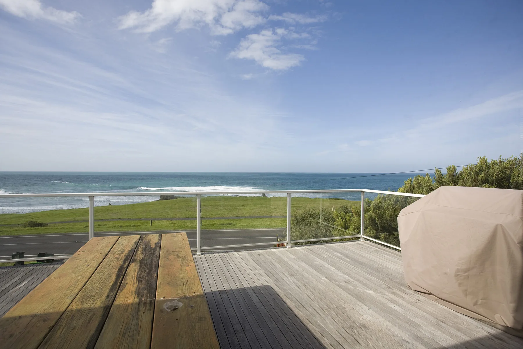 A wooden deck overlooks the ocean, with a glass railing and a covered outdoor item.