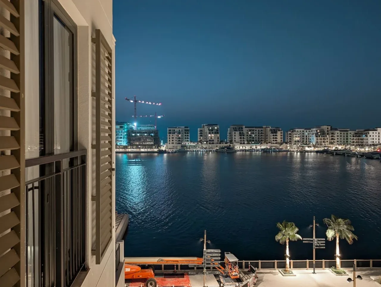 Vista nocturna desde un balcón hacia edificios iluminados y palmeras junto al agua.