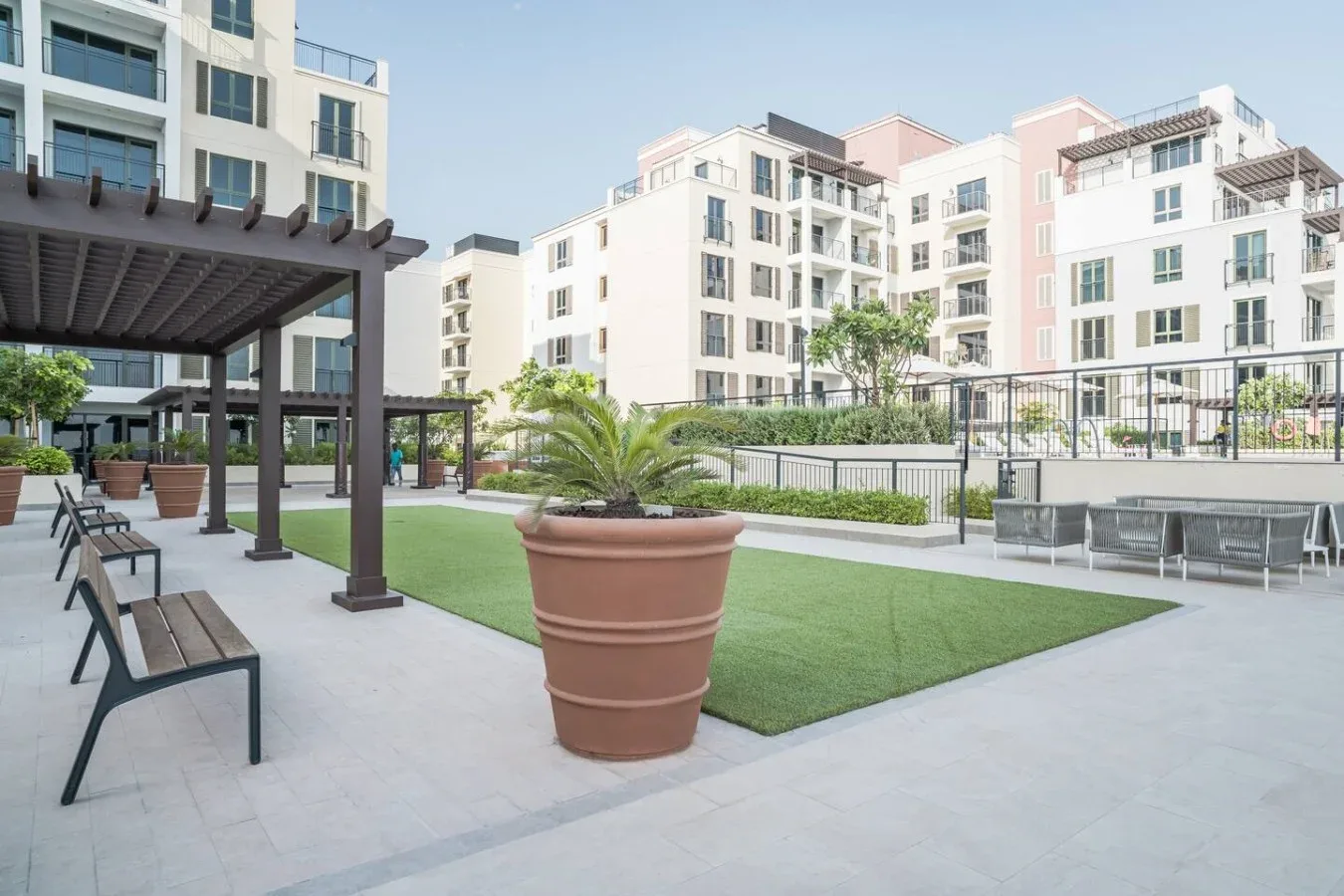 Un patio exterior tiene césped, plantas en macetas y asientos bajo una pérgola.