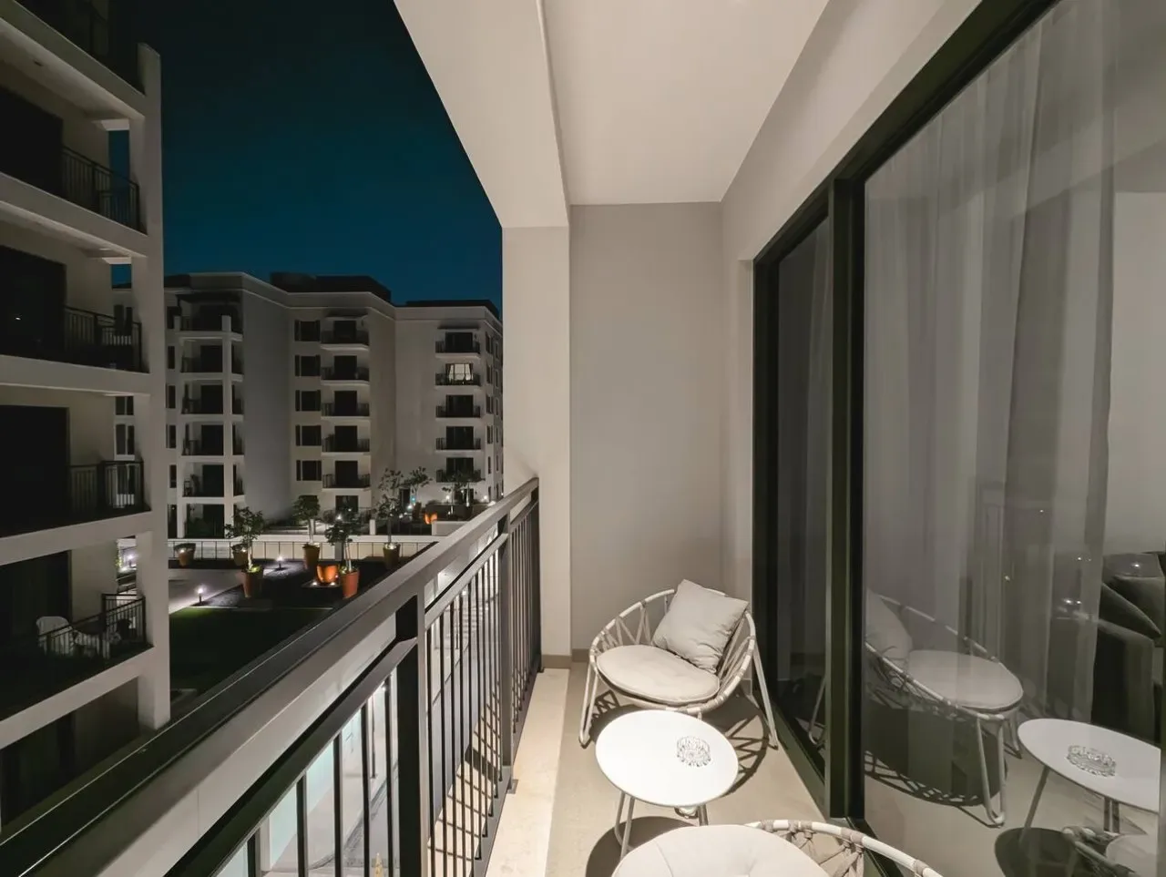 Un balcón estrecho de noche tiene asientos y una mesa pequeña junto a puertas corredizas de vidrio.