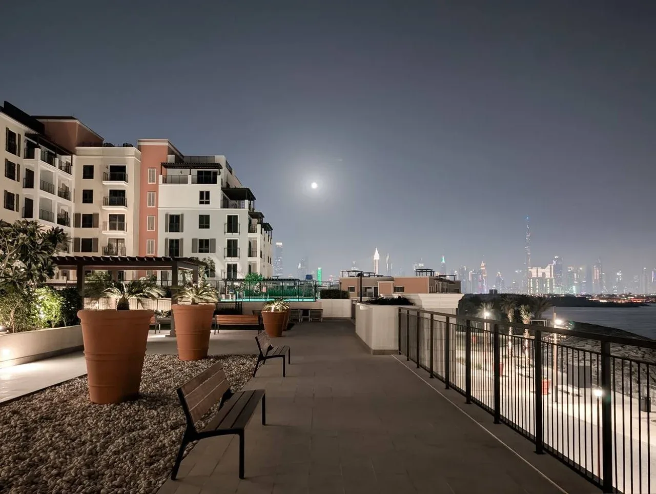 Una terraza en la azotea mira hacia la ciudad de noche, con jardineras y asientos junto al pasillo.