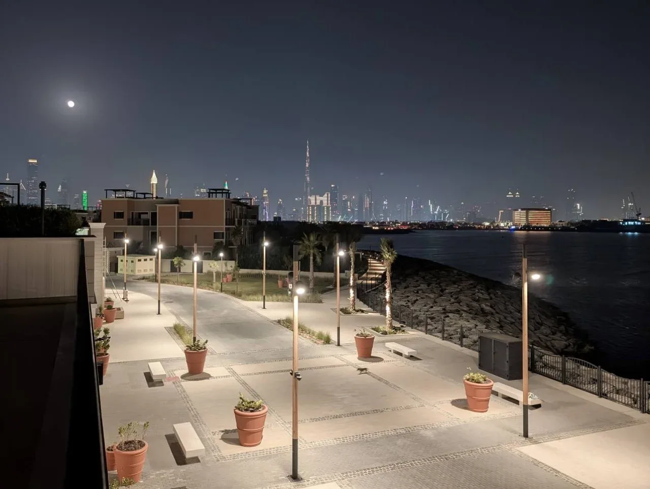 Un paseo junto al agua de noche, con pavimento iluminado y luces de la ciudad a lo lejos.