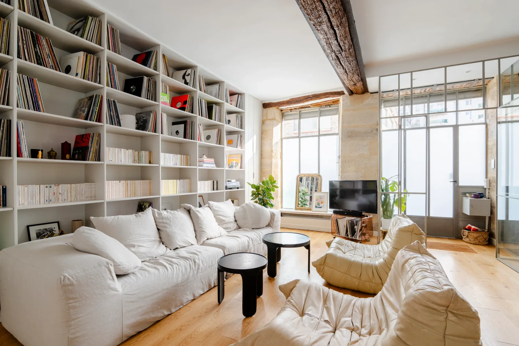 Una sala de estar luminosa con asientos blancos, estanterías y un televisor.