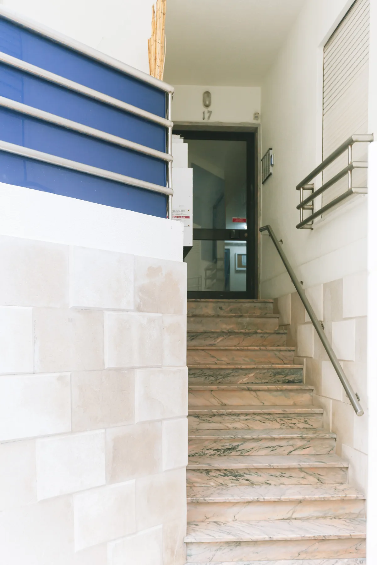 Una escalera interior con peldaños de mármol y pasamanos metálicos conduce a una entrada de vidrio.
