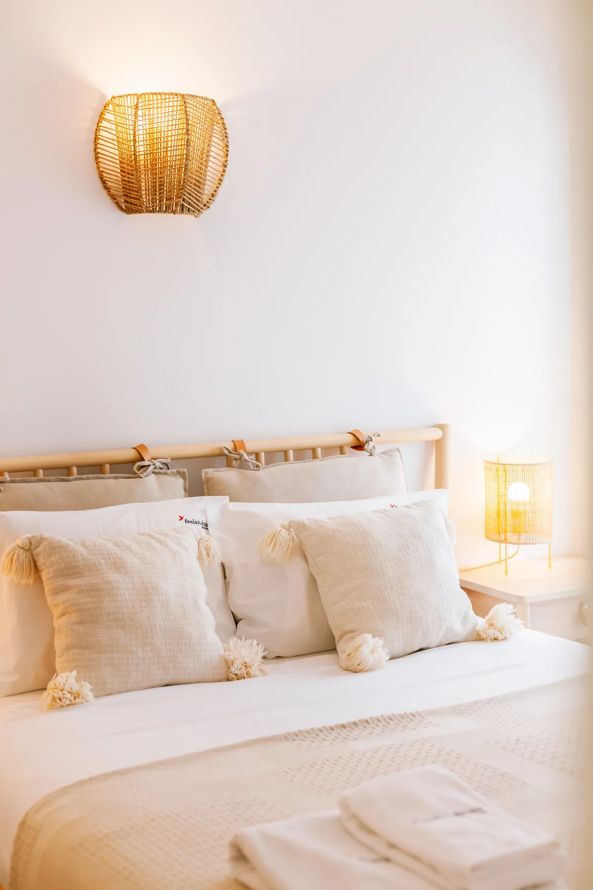 A bright bedroom with a bed, pale pillows, and warm woven lamps.