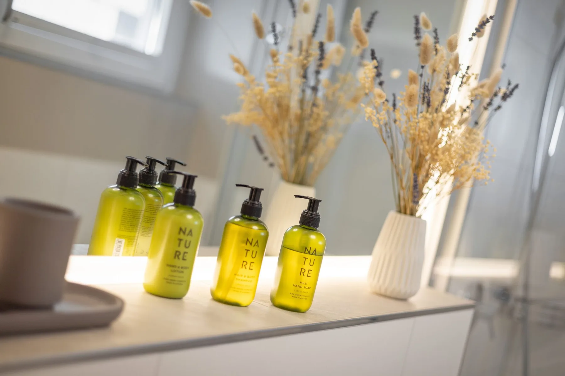 Tocador de baño con botellas amarillas con dosificador y flores secas en un jarrón.