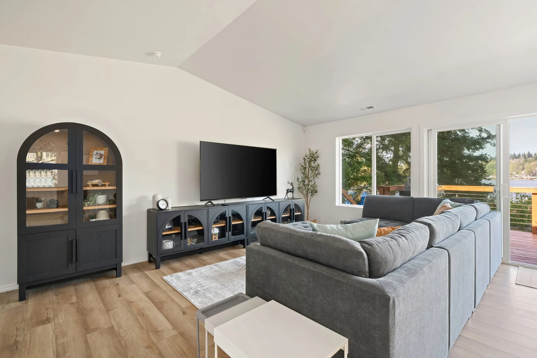 Una sala luminosa con asientos grises, un televisor y ventanas grandes.
