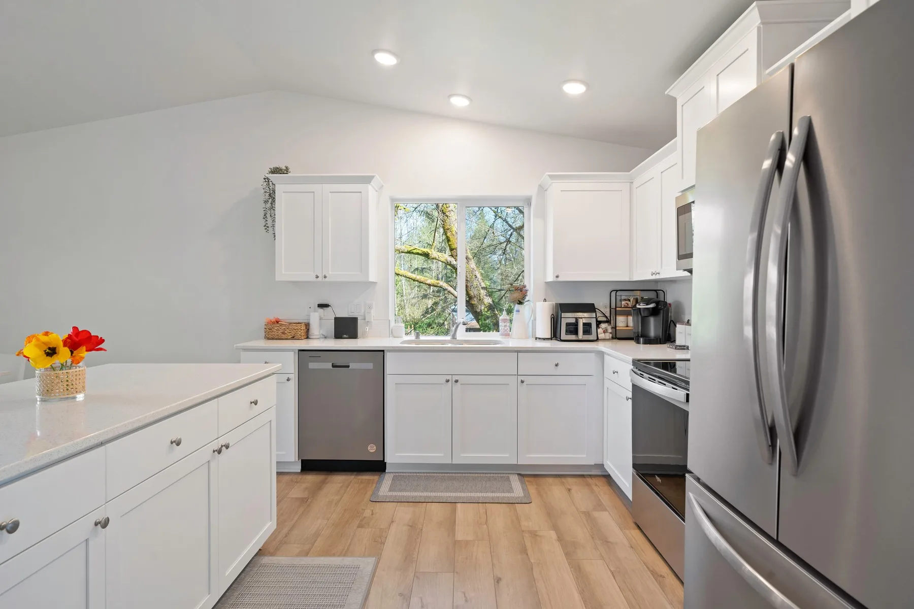Una cocina luminosa con gabinetes blancos, electrodomésticos de acero inoxidable y una ventana con vista a los árboles.