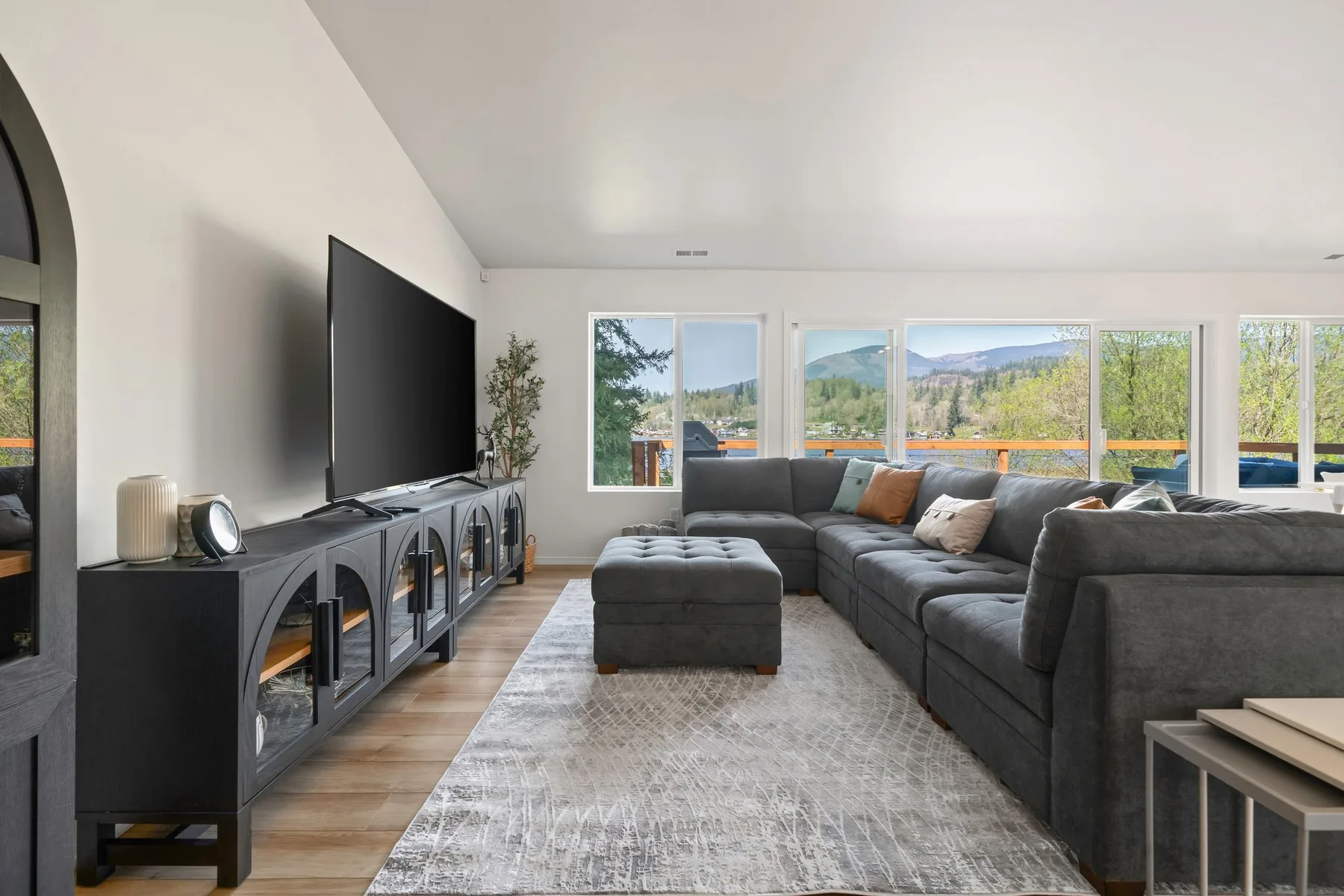 Una sala de estar con asientos grises, un televisor y amplias ventanas con vistas a árboles y colinas.