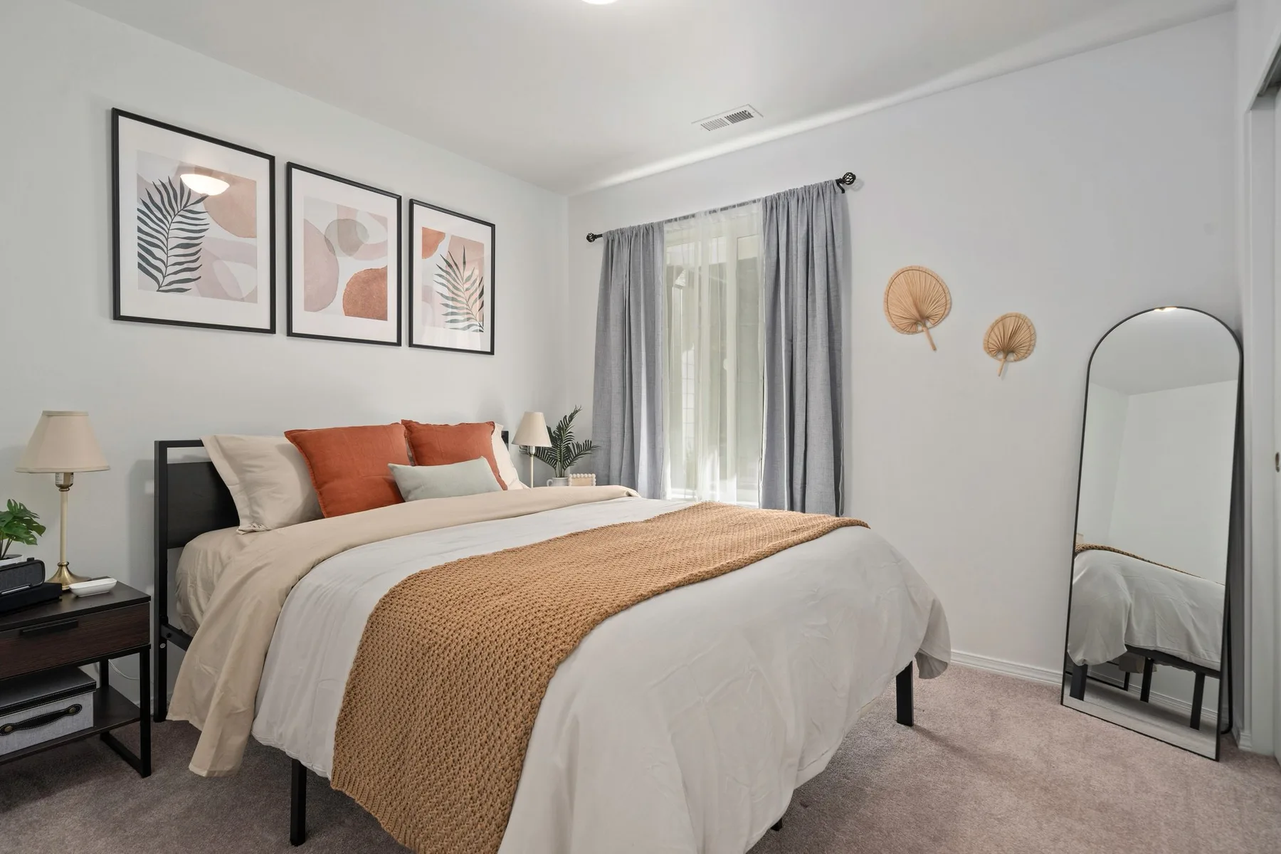 Un dormitorio luminoso con una cama tendida, láminas enmarcadas y ventanas con cortinas.