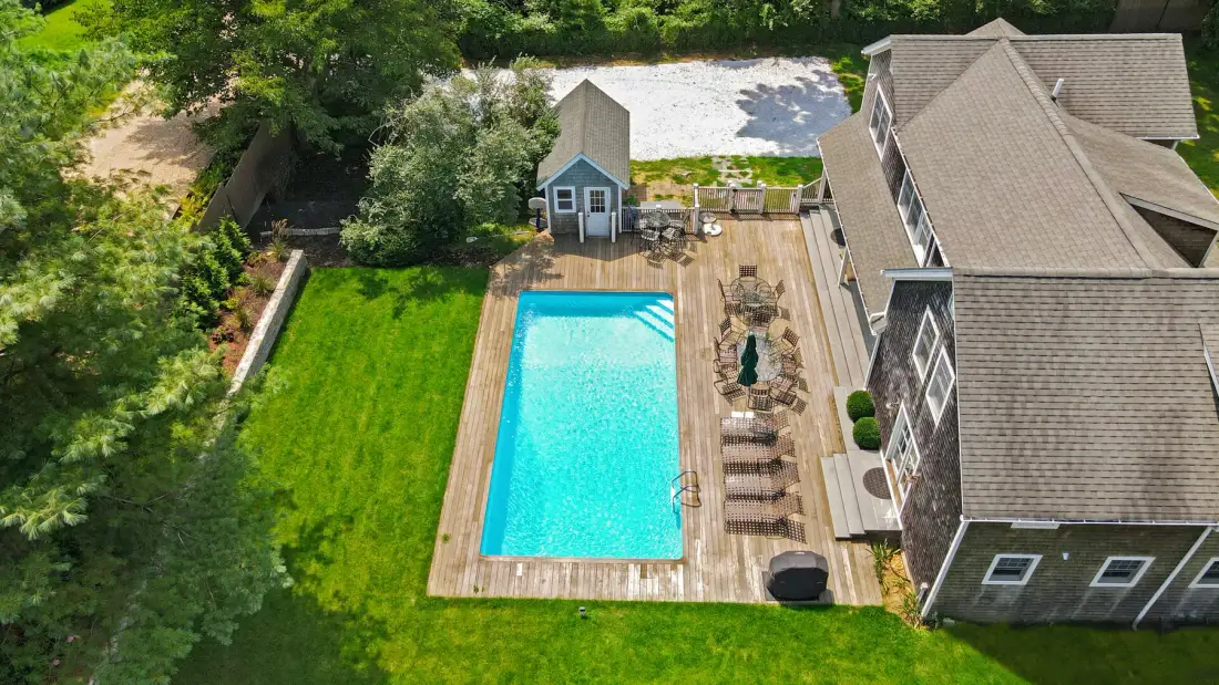 An aerial view of a backyard pool beside a house, surrounded by lawn and trees.