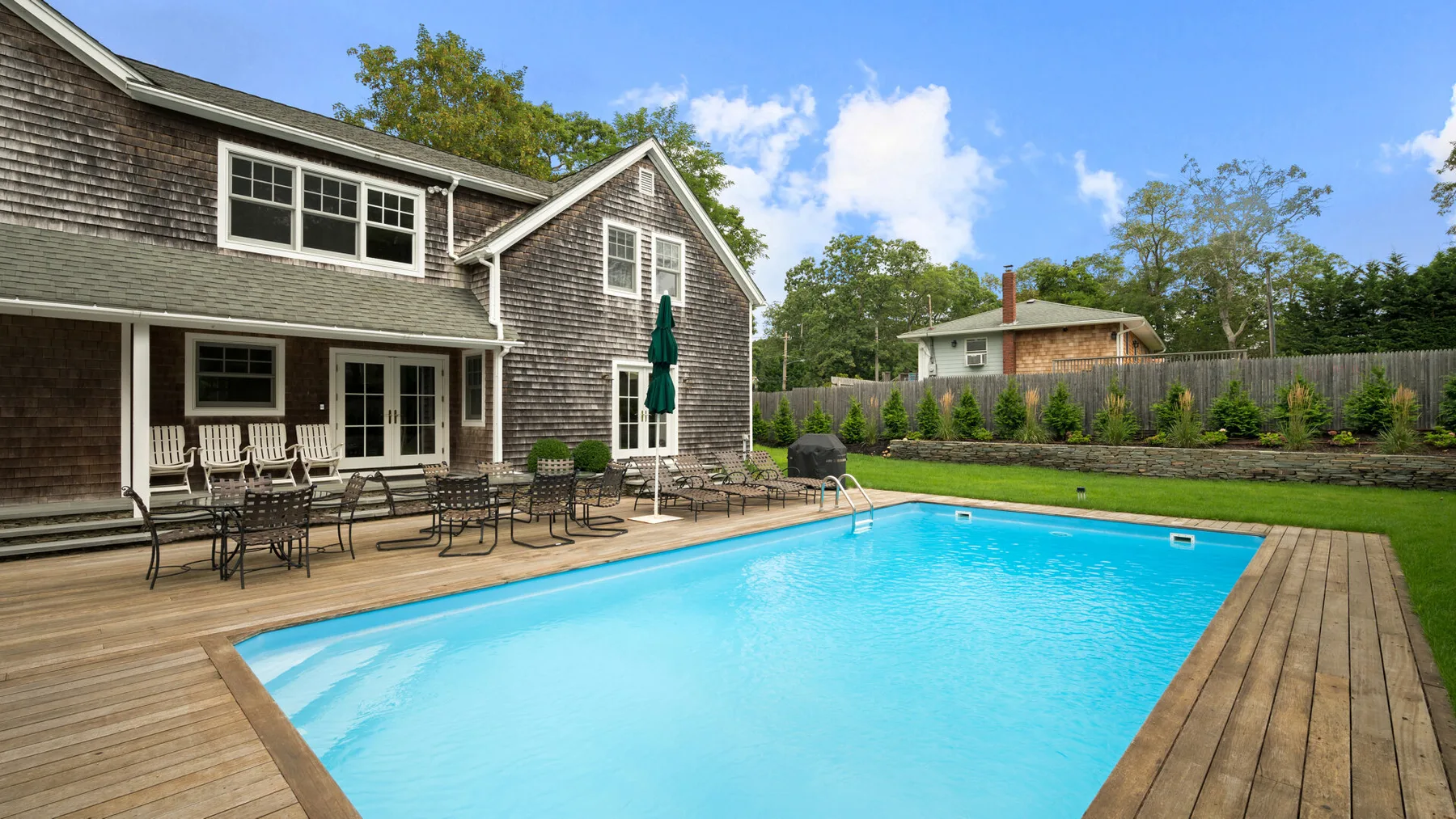 An outdoor swimming pool sits beside a wooden deck and a shingle-sided house.