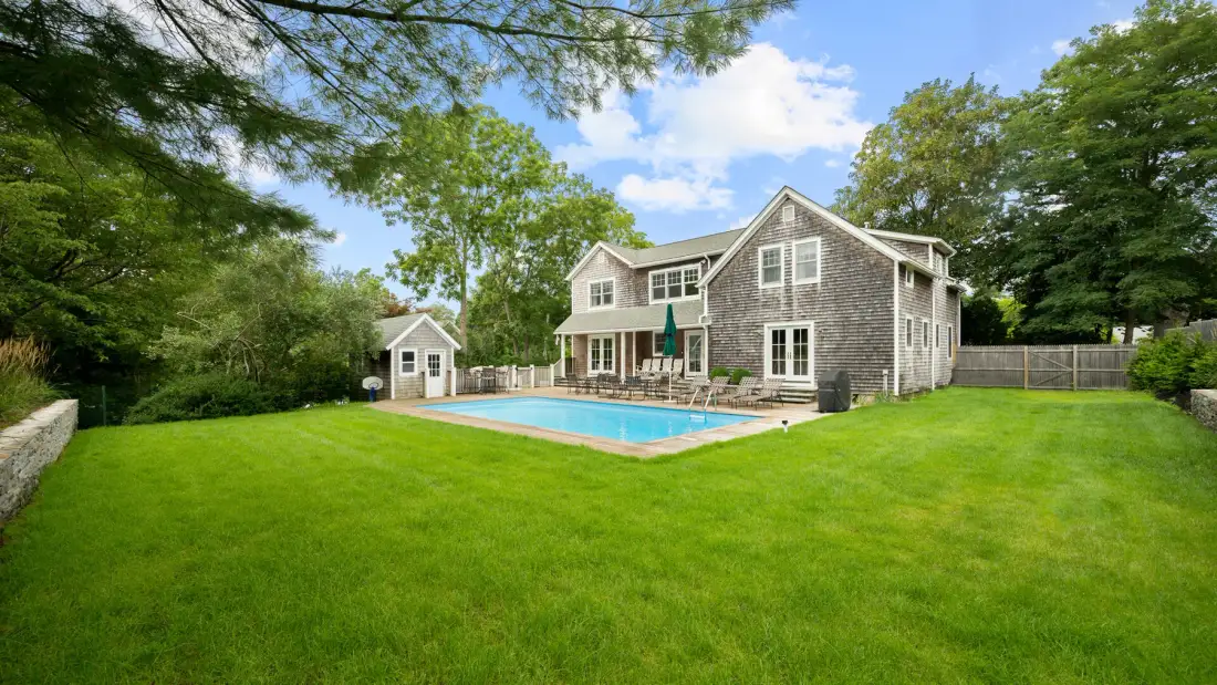 A large house stands beside a swimming pool in a grassy yard surrounded by trees.