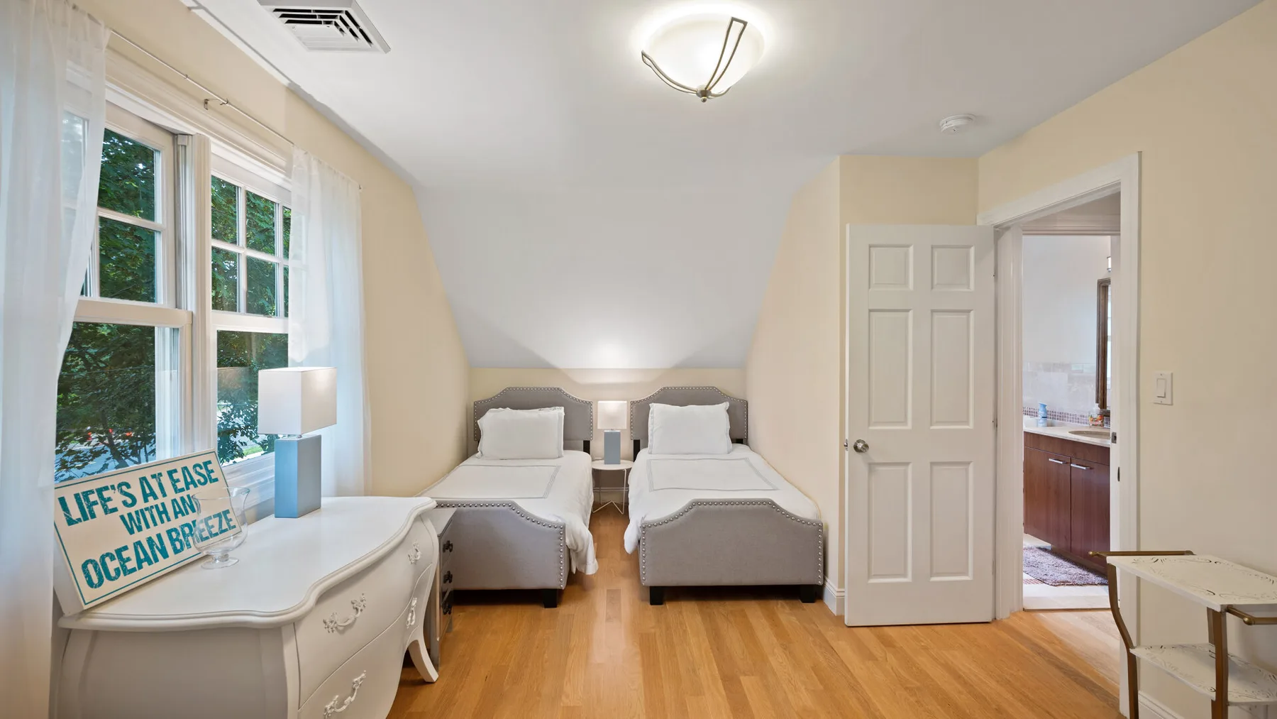 A bright bedroom with two beds, large windows, and an open doorway.